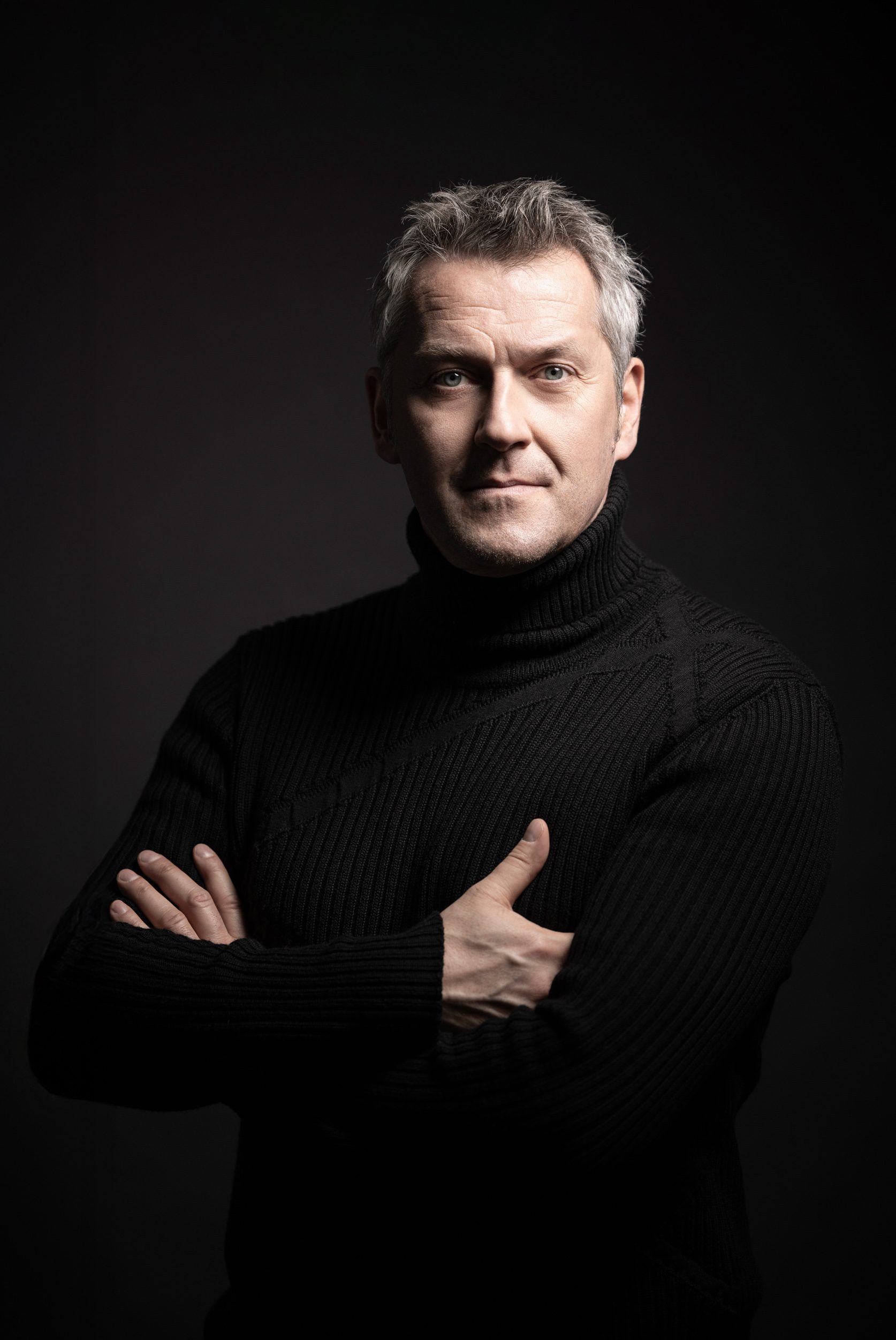A middle-aged man with short, light gray hair stands confidently with his arms crossed, wearing a black turtleneck sweater. He has a serious expression, set against a dark background that enhances his features. The image conveys a sense of strength and purpose.
