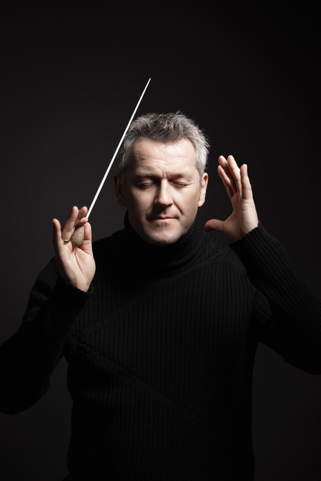 A man in a black turtleneck is conducting music, holding a conductor's baton in one hand. His eyes are closed, and he appears focused and expressive, surrounded by a dark background.