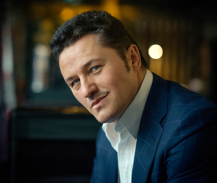 A man in a blue suit with a checkered pattern is seated, looking slightly to his right with a thoughtful expression. He has short, dark hair and is wearing a white shirt underneath his suit jacket. The background is softly blurred.