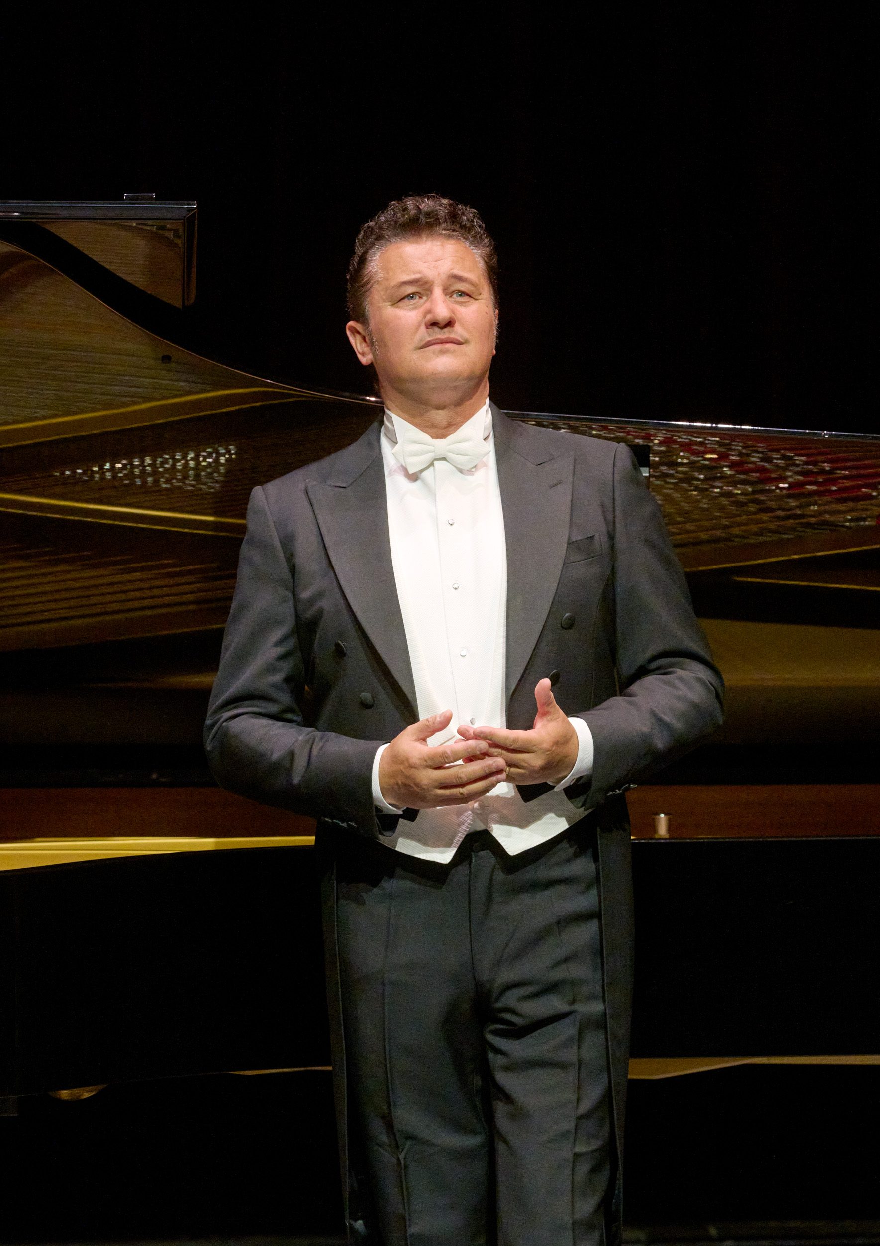A man wearing a formal black tuxedo stands with his hands clasped in front of him, looking thoughtfully into the distance. He is positioned on a stage, with a grand piano visible behind him, suggesting a musical performance setting.