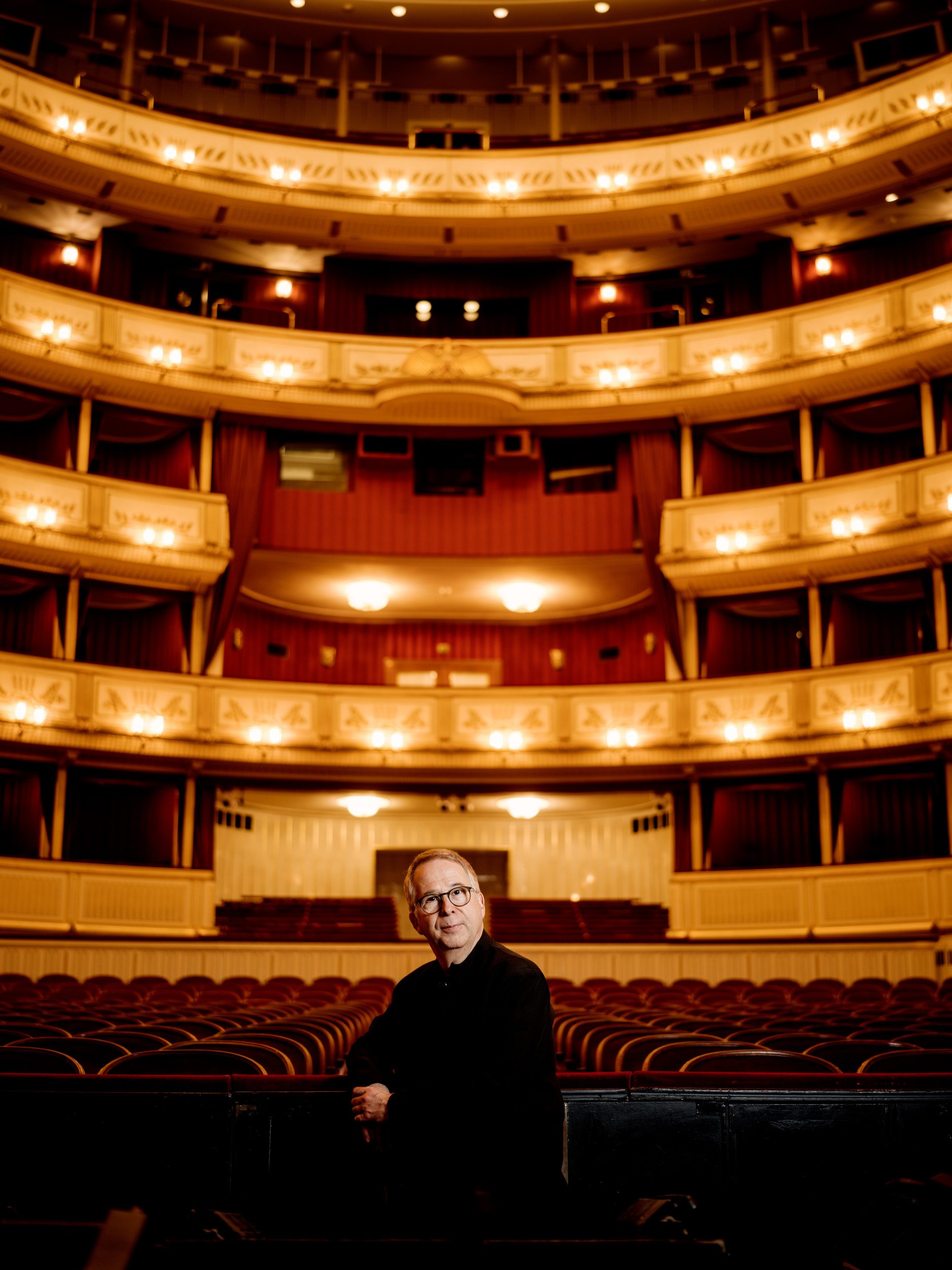 A middle-aged man with glasses is seated in an empty theater, gazing thoughtfully. The elegant interior features tiered seating and warm lighting, creating a rich atmosphere. The stage is dimly lit, suggesting a sense of anticipation for an upcoming performance.