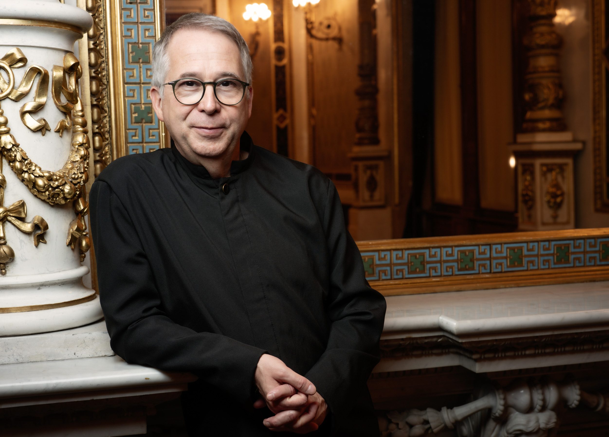 A middle-aged man wearing glasses and a black outfit stands confidently beside a decorative column. He is smiling slightly, with his arms crossed, in an elegantly styled setting that suggests an artistic or historic venue.  The background features intricate architectural details.