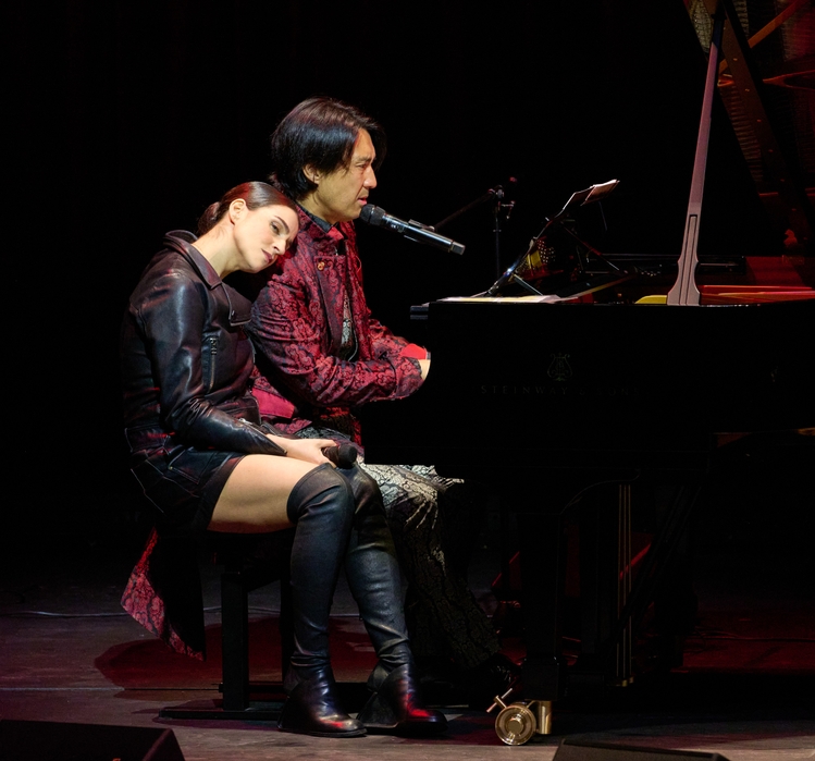 Ein Musiker sitzt am Piano und singt, während eine Frau mit sanfter Pose an ihn gelehnt ist. Beide tragen elegante Kleidung und scheinen in eine emotionale Darbietung vertieft zu sein, umgeben von einer dunklen Bühnenatmosphäre.