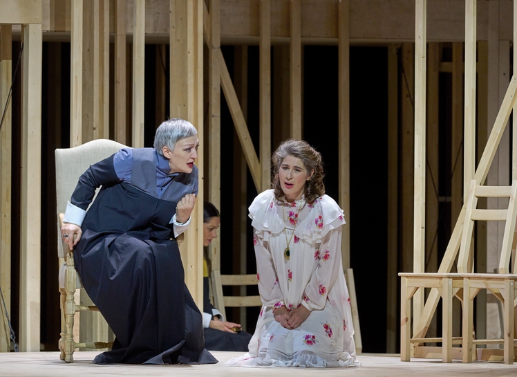 Zwei Frauen sprechen vor einem Holzgerüst; die eine kniet auf dem Boden, die andere beugt sich zu ihr herunter.