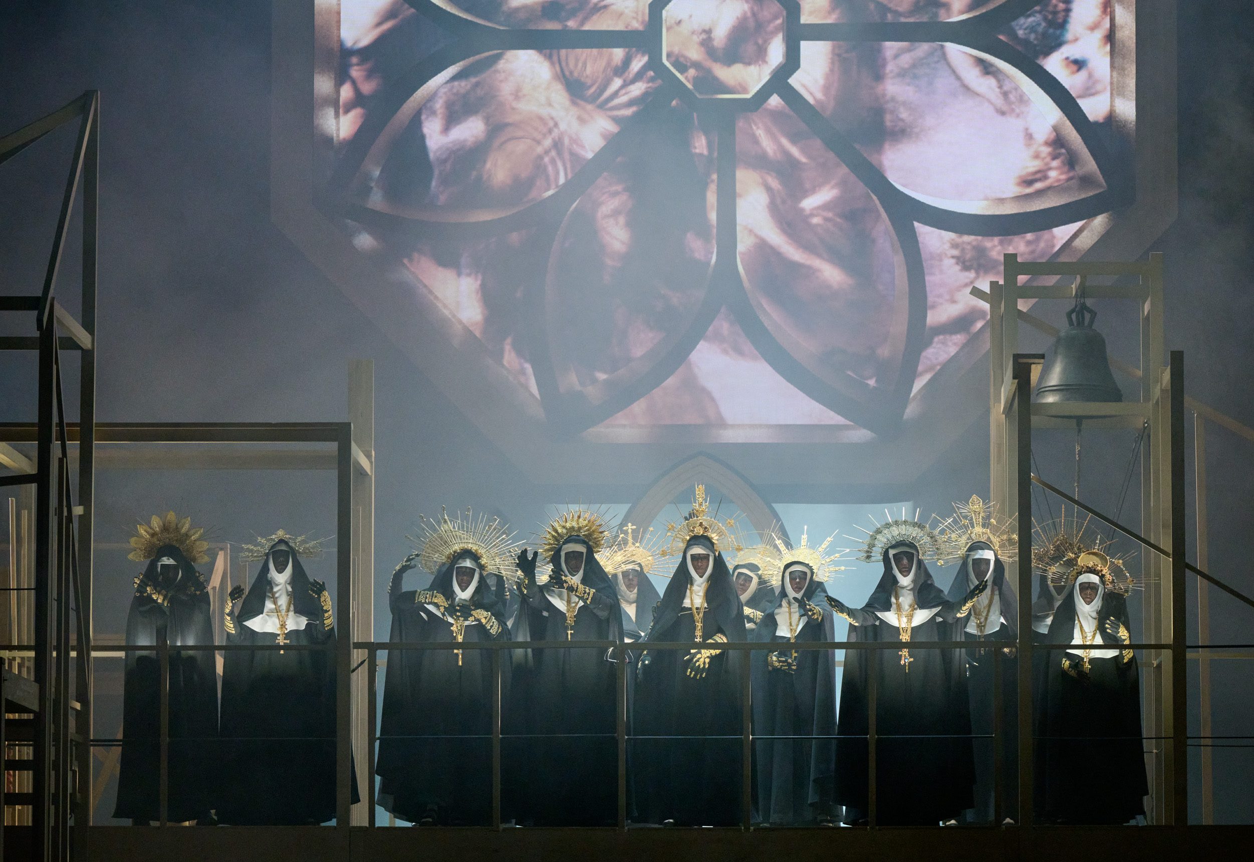 Mehrere dunkel gekleidete Gestalten mit goldenen Kopfbedeckungen stehen unter einem großen Kirchenfenster auf einer eingenebelten Bühne.