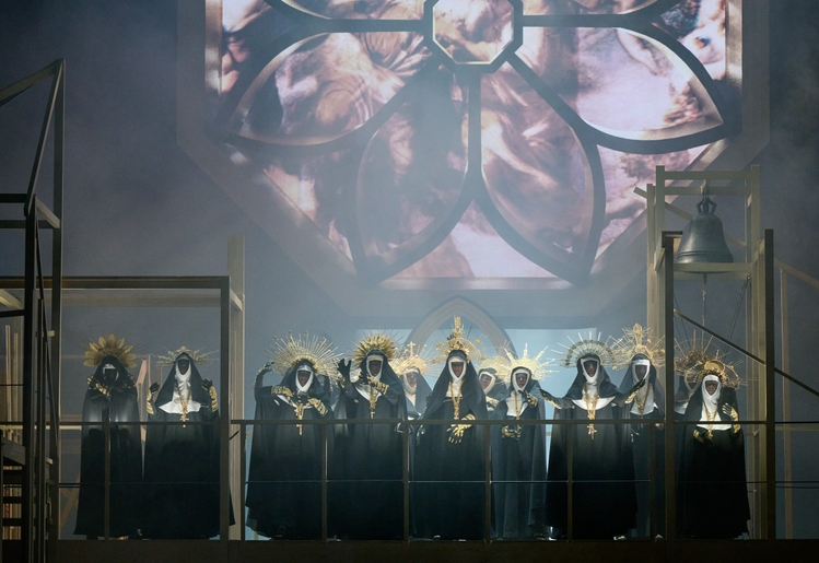Mehrere dunkel gekleidete Gestalten mit goldenen Kopfbedeckungen stehen unter einem großen Kirchenfenster auf einer eingenebelten Bühne.