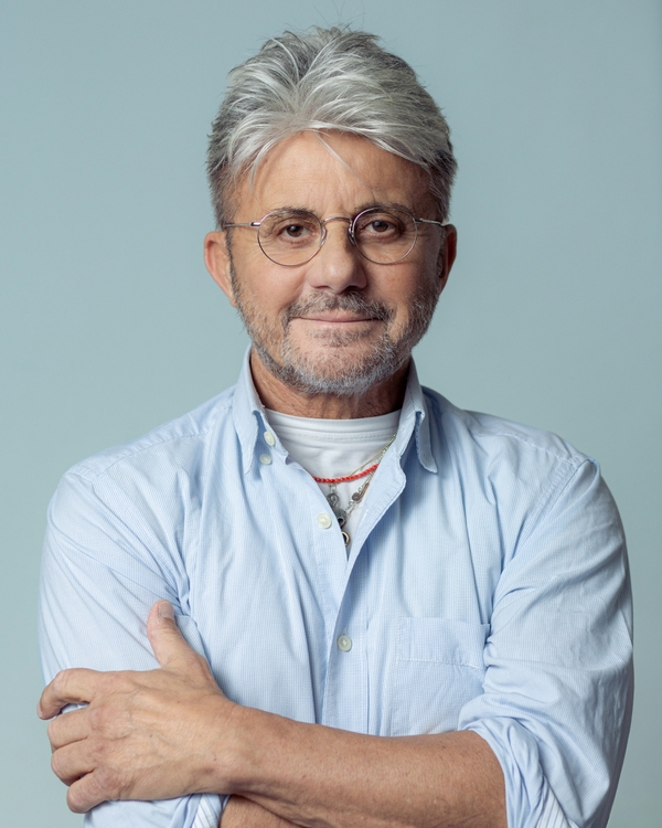 A middle-aged man with light gray hair and wearing round glasses smiles gently. He is dressed in a light blue button-up shirt and crosses his arms. The background is a soft, neutral color, emphasizing his friendly demeanor.