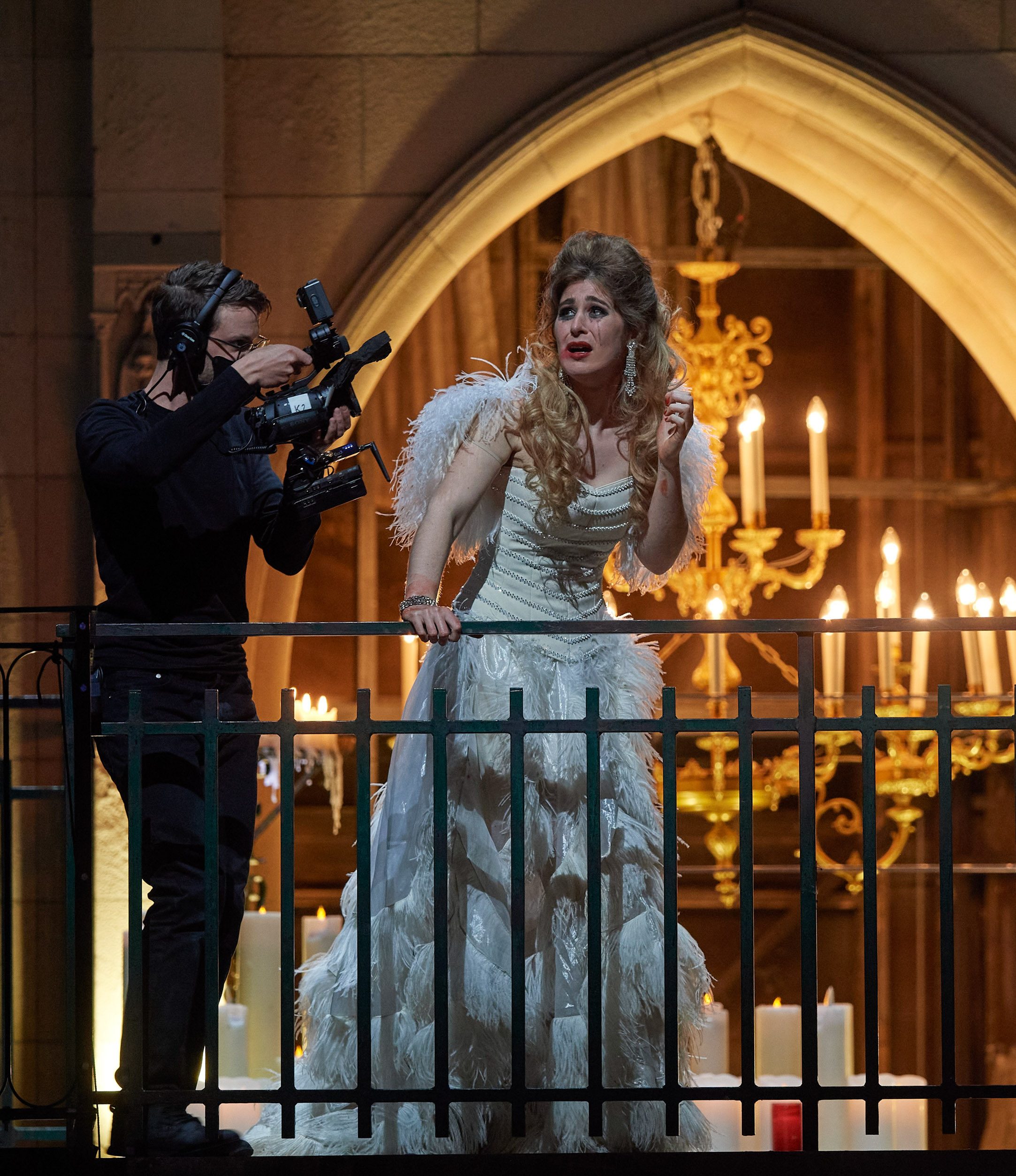Eine Frau in weißem Kleid blickt erschrocken nach vorne und hält sich dabei an einem Geländer fest. Sie wird währenddessen von einer schwarz gekleideten Kameraperson gefilmt.