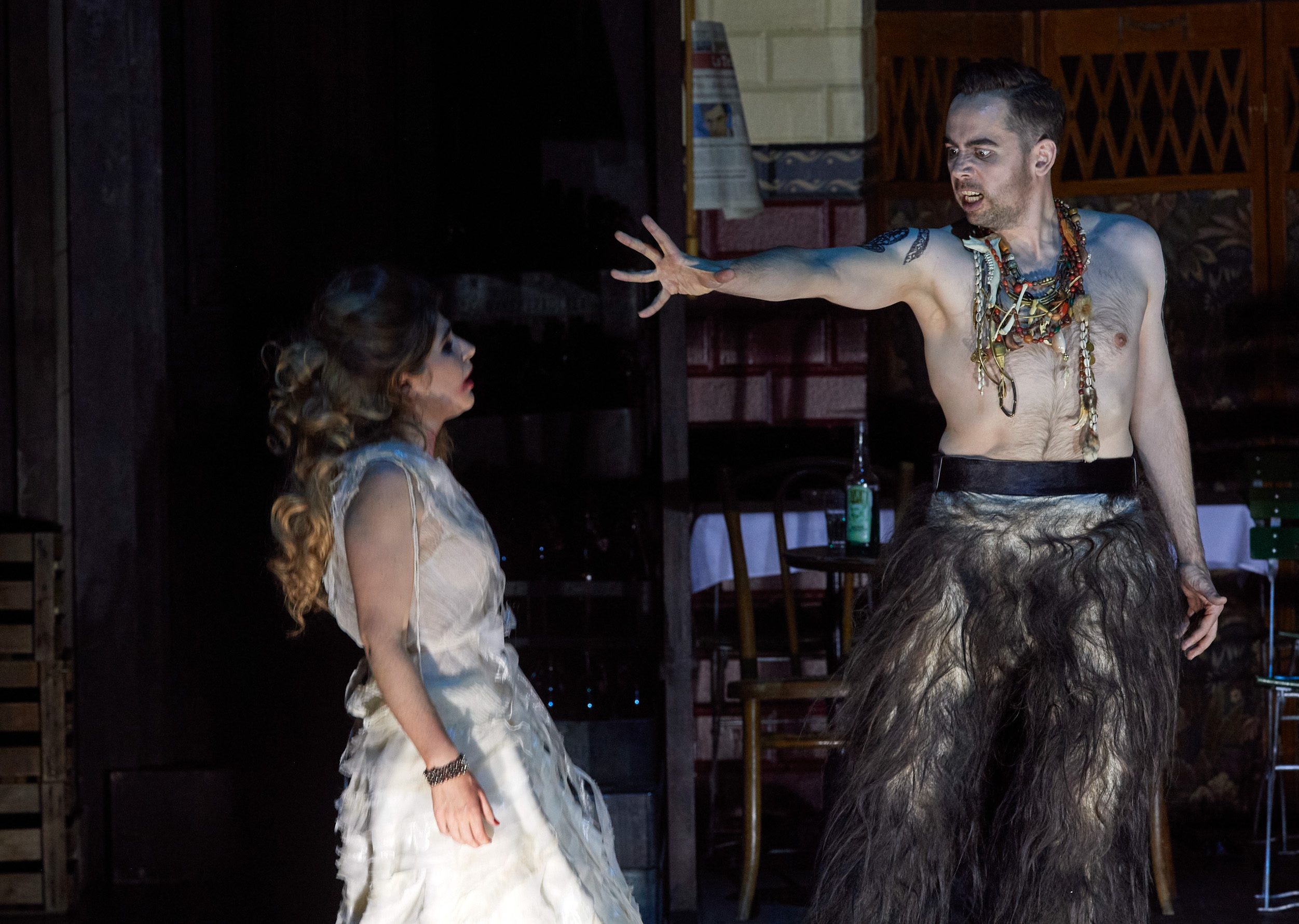 Ein furchteinflößender Mann mit nacktem Oberkörper streckt seine Hand in die Richtung einer Frau in weißem Kleid.