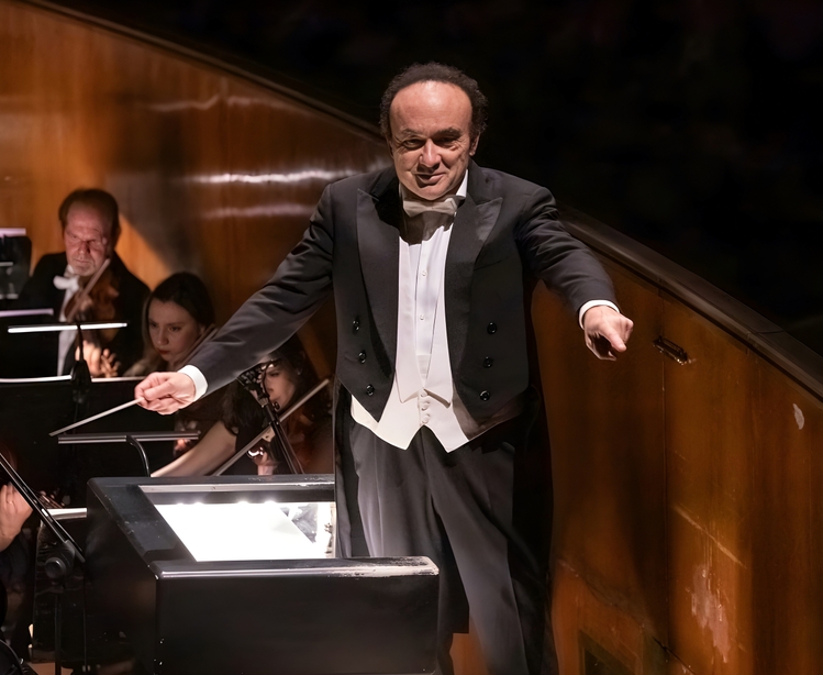 Ein Dirigent in schwarzem Frack steht in einem Orchestergraben und zeigt zur Seite