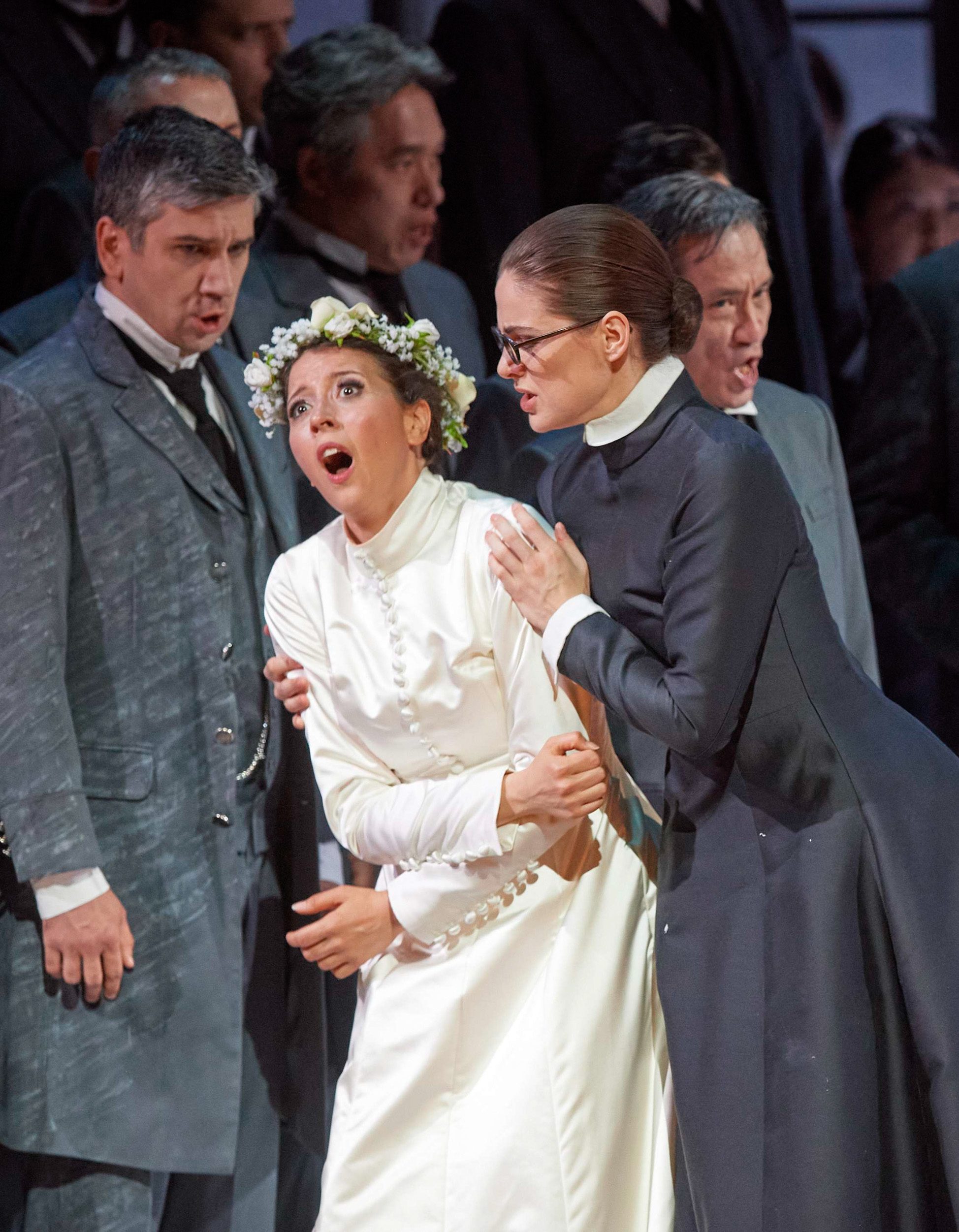 Eine Frau im weißen Kleid und mit einem Blumenkranz auf dem Kopf wird von einer dunkel gekleideten Frau gehalten.