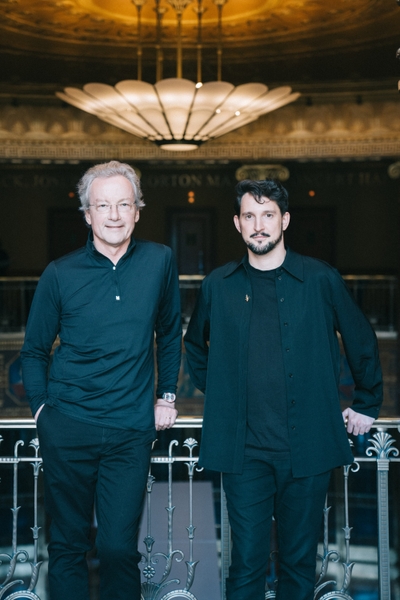 Zwei Männer stehen nebeneinander an einem Geländer in einem eleganten Raum. Beide tragen schwarze Kleidung. Im Hintergrund ist eine kunstvolle Decke sowie eine große Lampe zu sehen, die eine warme Atmosphäre schafft.