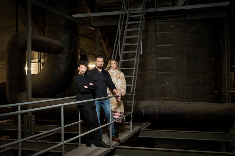 Drei Personen stehen auf einer Plattform in einem industriellen Raum. Die Frau in einem langen, gemusterten Mantel steht rechts, während zwei Männer in dunkler Kleidung, einer mit verschränkten Armen, in der Mitte und links stehen. Im Hintergrund sind große, dunkle Rohre sichtbar.