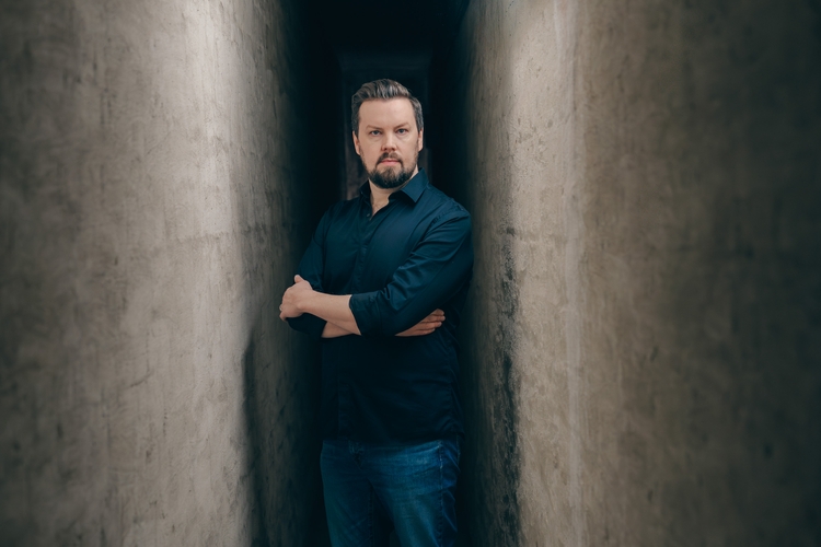 Ein Mann mit Bart steht in einem engen, schmalen Gang zwischen zwei Betonwänden. Er trägt ein schwarzes Hemd und Jeans, hat die Arme vor der Brust verschränkt und schaut ernst in die Kamera. Das Licht ist gedämpft, was eine nachdenkliche Atmosphäre erzeugt.