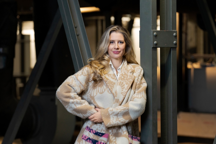 Eine Frau mit langen, lockigen Haaren steht an einer Metallstruktur. Sie trägt einen beigefarbenen, gemusterten Mantel und hat ihre Hände in der Nähe ihrer Taille verschränkt. Ihr Blick ist freundlich, während sie in die Kamera schaut.