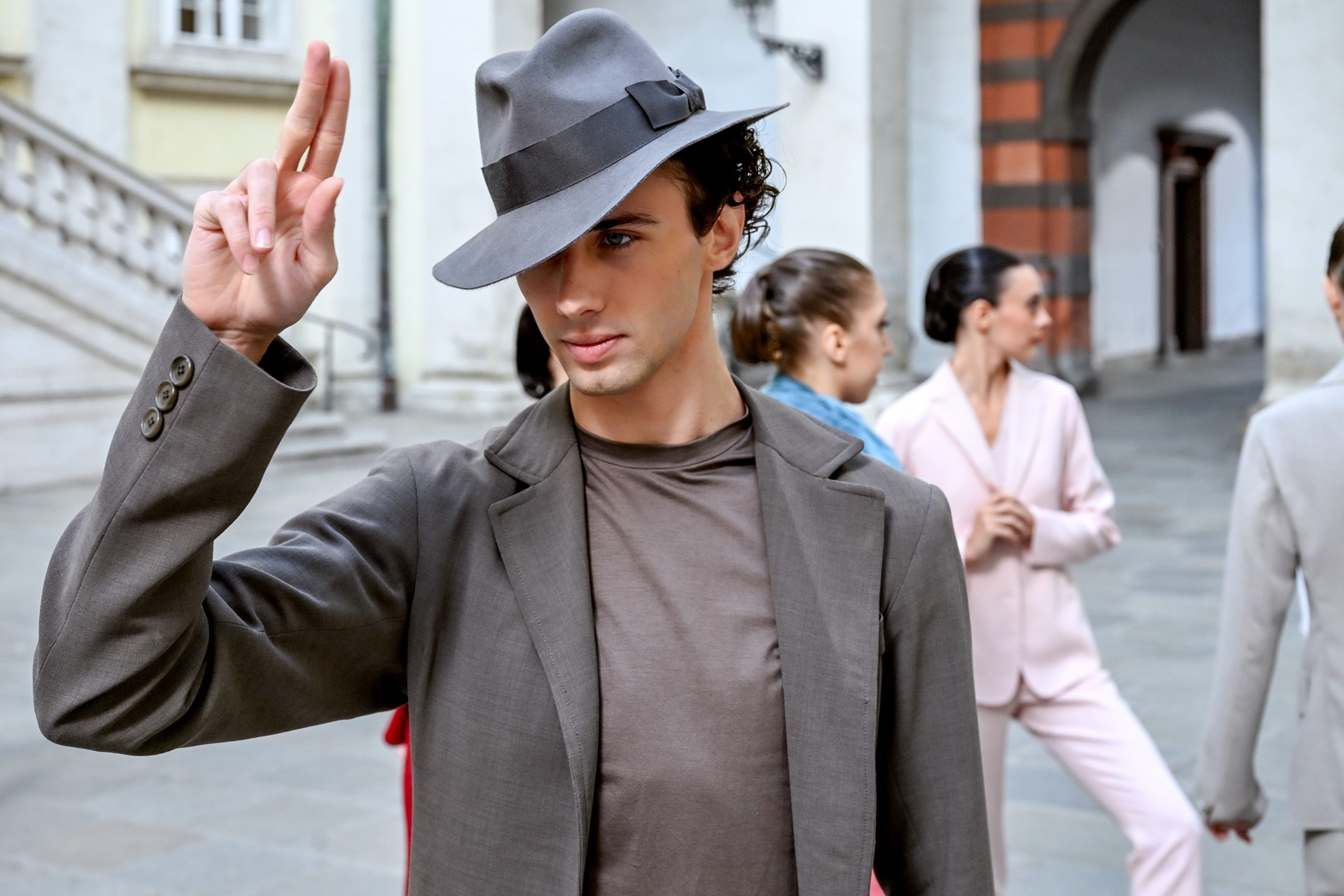 Ein junger Mann in einem grauen Anzug und Hut macht eine Geste mit seiner Hand. Im Hintergrund sind weitere Personen in Modeoutfits zu sehen. Die Szene scheint in einer urbanen Umgebung zu spielen, möglicherweise in einem Innenhof oder auf einem Platz.