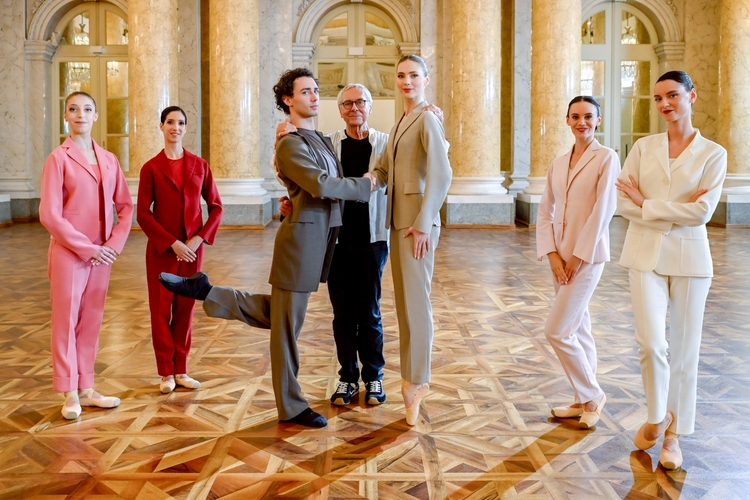 Sechs Tänzer und Tänzerinnen stehen in eleganten Anzügen in einem großen, heller Raum mit einem Holzboden. Ein Tänzer hebt eine Trainerfigur in der Mitte hocherhoben und lächelt. Die Atmosphäre ist kreativ und professionell.