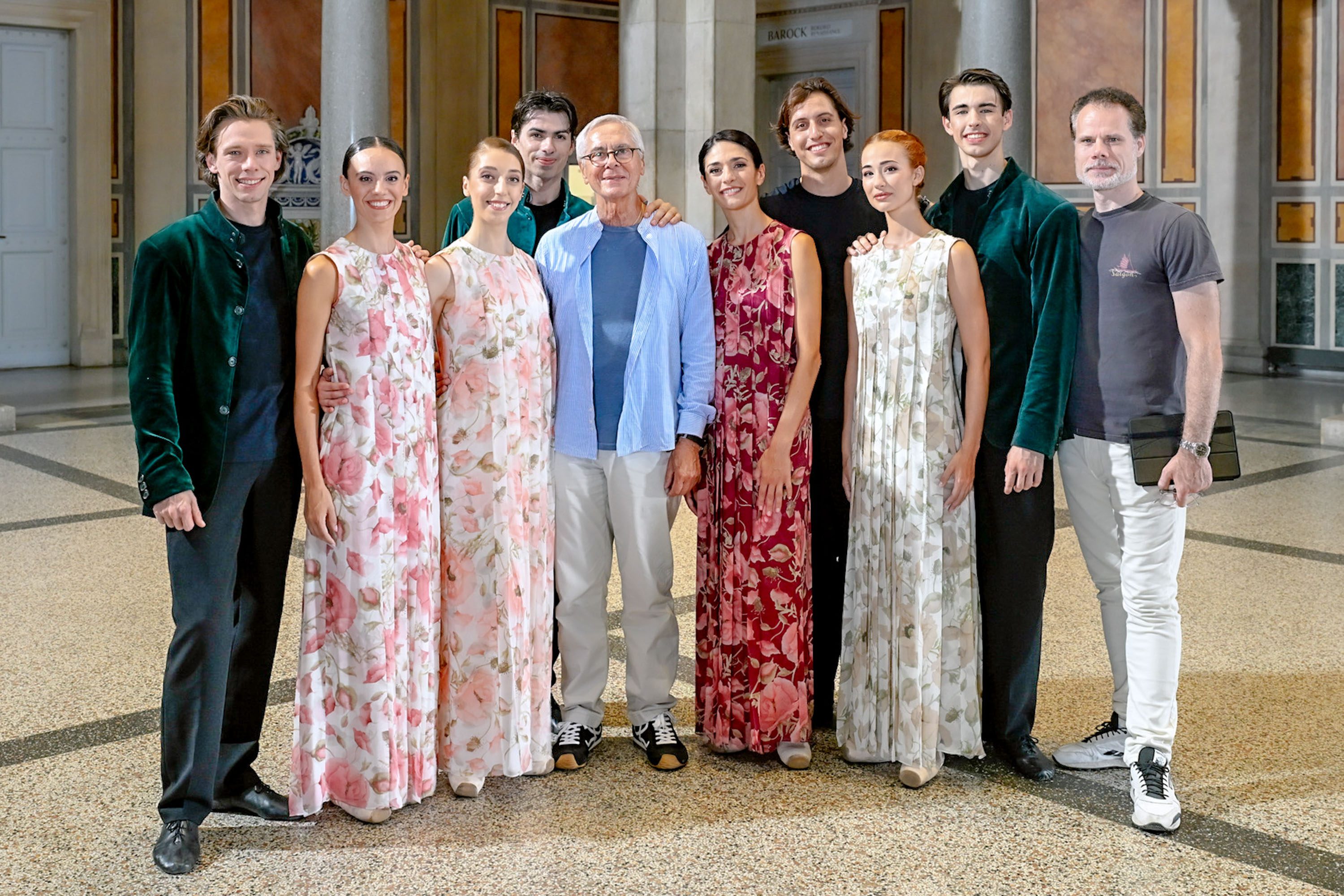 Eine Gruppe von acht Personen steht im Innenraum eines Gebäudes. Sie tragen elegante Kleidung mit floralen Mustern. In der Mitte steht ein älterer Mann mit einer leichten Bluse. Die Stimmung ist freundlich und aufgeschlossen.
