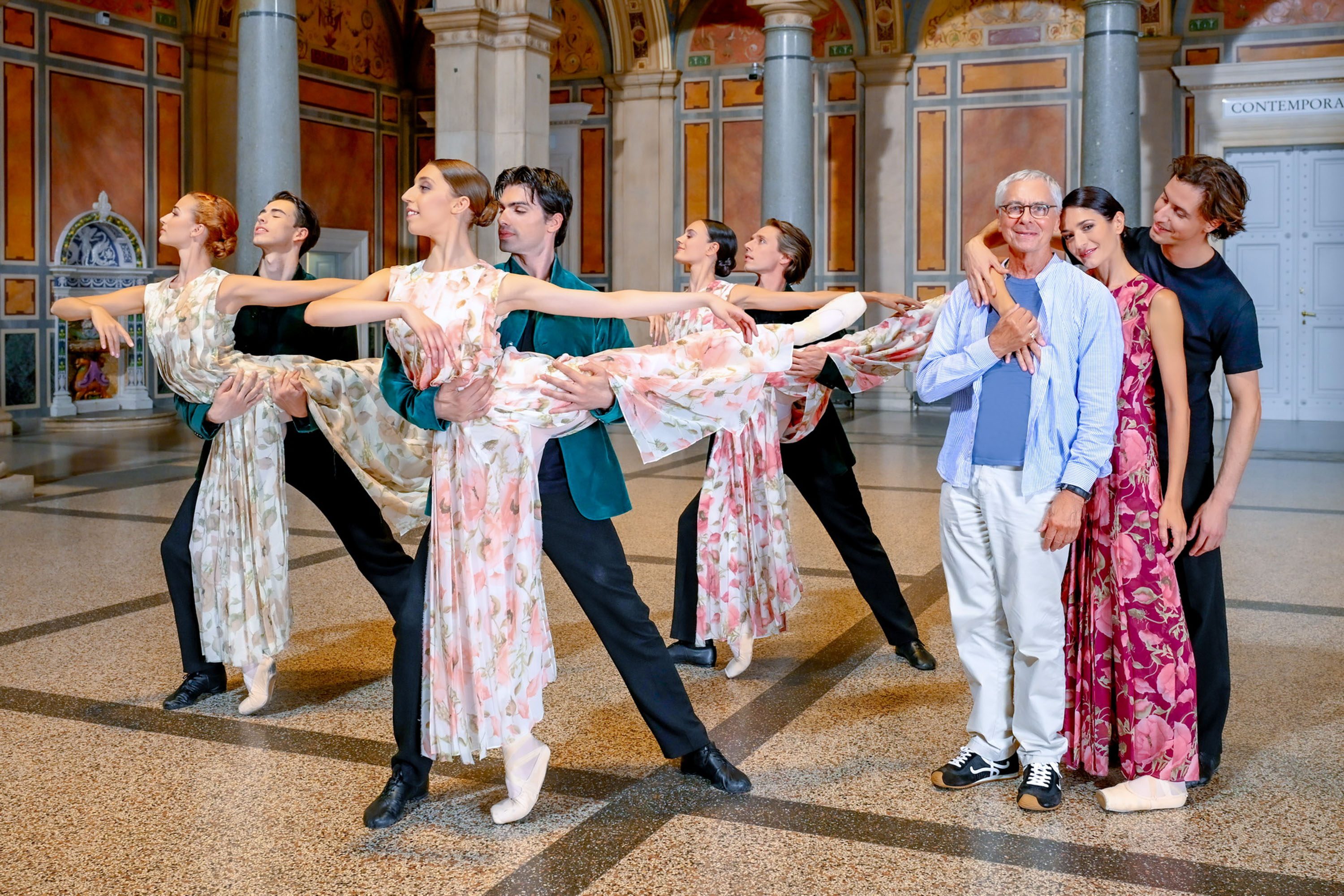Eine Gruppe von fünf Tänzern in floralen Kostümen führt eine elegante Tanzpose aus. Im Vordergrund stehen zwei Personen, die lächeln, während sie eine Verbindung zu den Tänzern zeigen. Die Umgebung ist architektonisch gestaltet mit bunten Wänden und einem großen Raum.