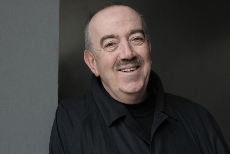 Ein lächelnder Mann mit grauem Haar und einem Schnurrbart trägt einen schwarzen Mantel. Er lehnt sich an eine graue Wand und blickt freundlich in die Kamera. Der Hintergrund ist dunkel und schafft einen Kontrast zu seinem Gesicht.