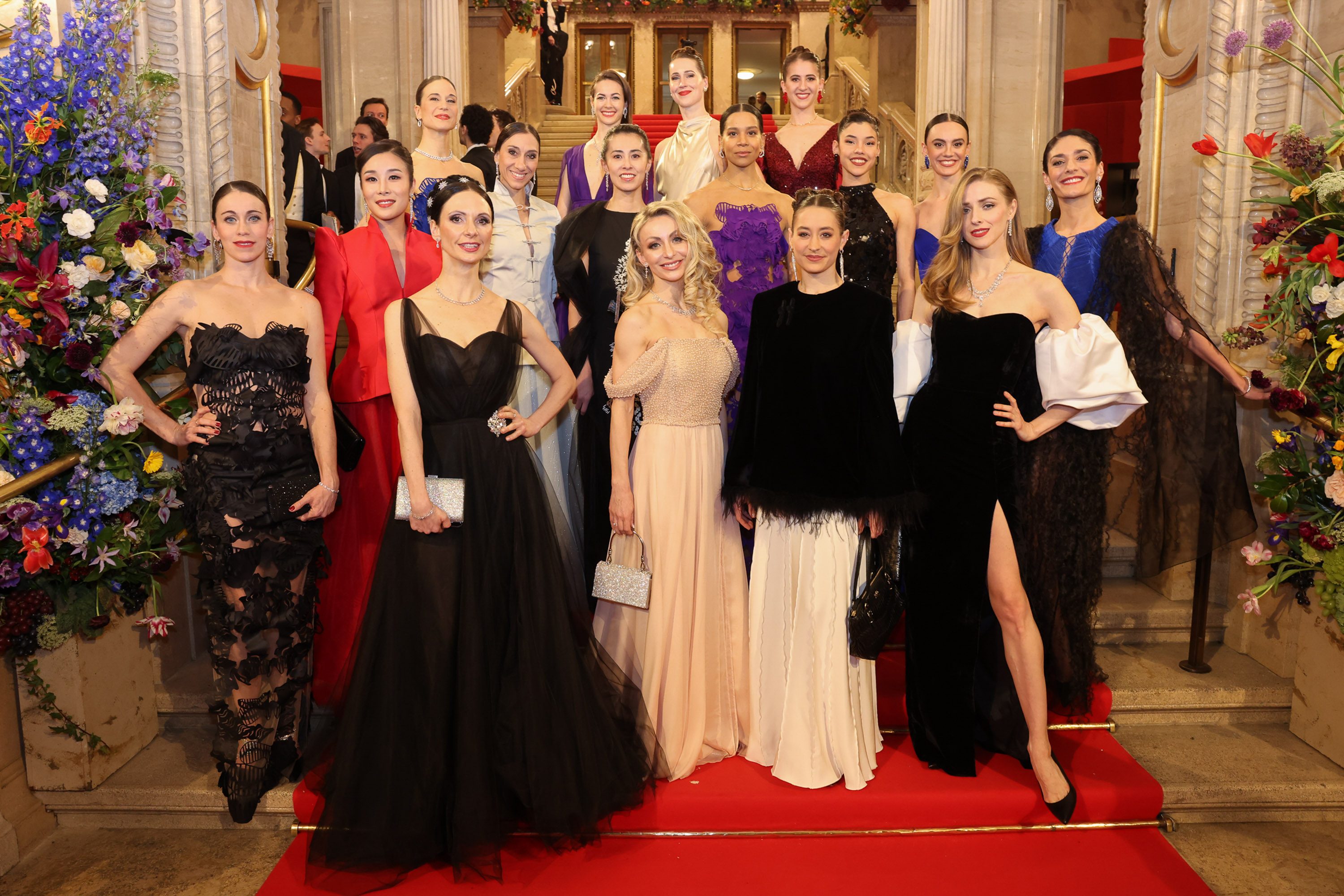 Eine Gruppe von 16 Frauen in eleganten Abendkleidern posiert auf einer roten Treppe. Sie stehen vor einer dekorativen Blumeninstallation. Das Ambiente ist festlich und glamourös, was die Feierlichkeit des Anlasses unterstreicht.