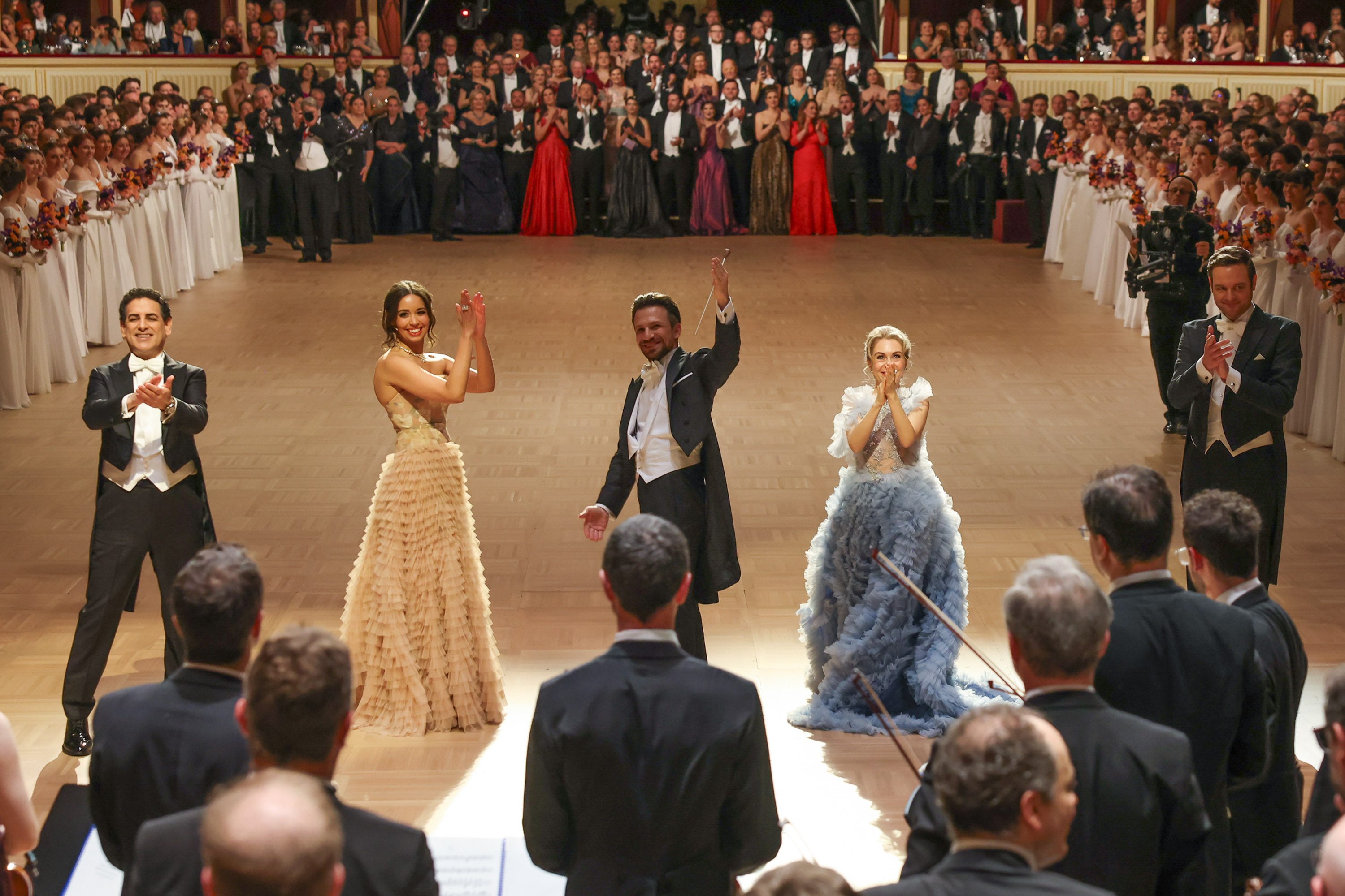 Auf einer festlichen Bühne stehen vier Künstler in eleganter Kleidung und applaudieren. Im Hintergrund applaudiert ein Publikum, das in formeller Abendgarderobe gekleidet ist. Die Atmosphäre ist festlich und feierlich, mit einem Gefühl von Begeisterung und Anerkennung.