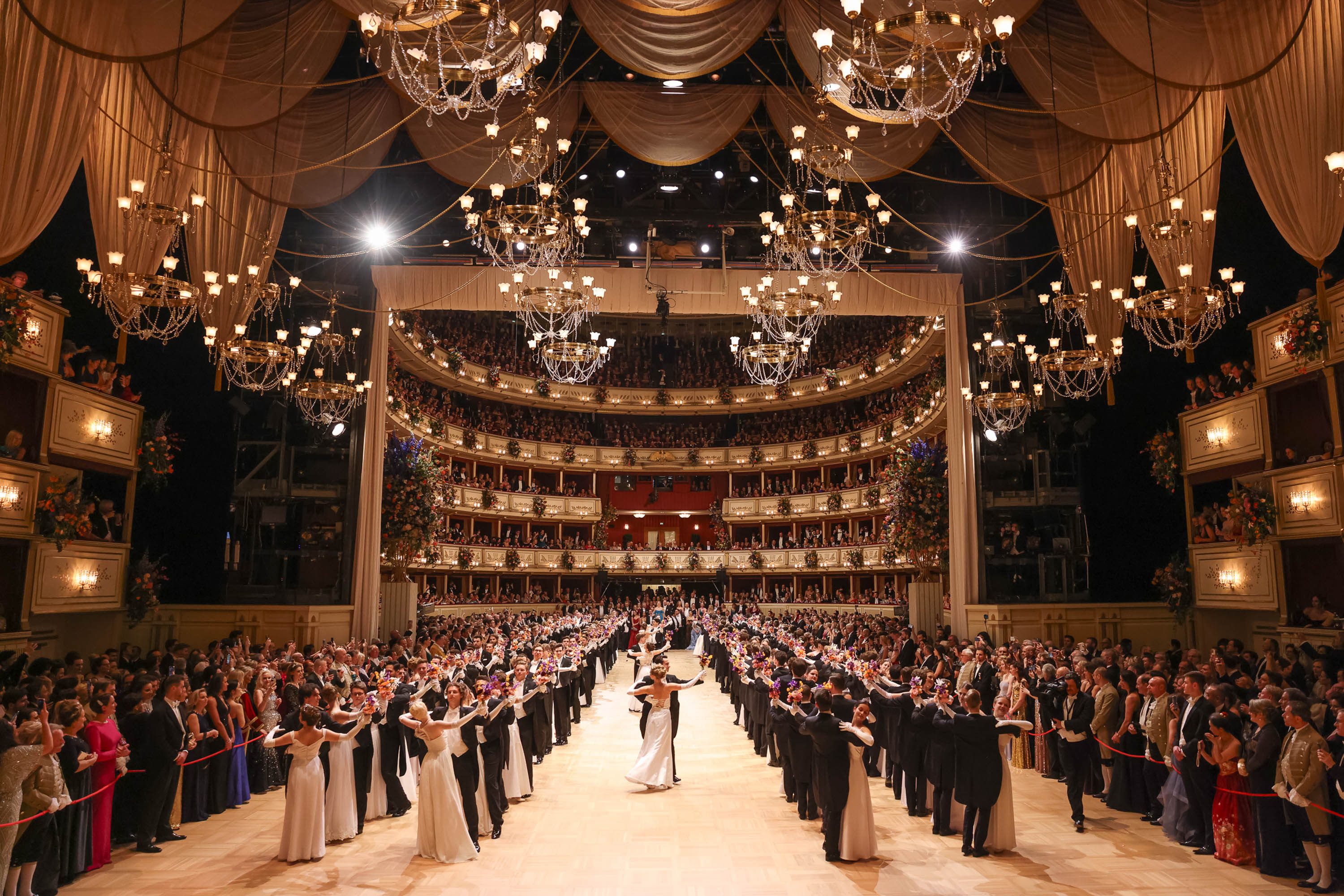 In einem festlichen Saal tanzen Paare elegant, umgeben von Zuschauern, die auf mehreren Etagen sitzen. Die aufwändige Beleuchtung und die Dekoration schaffen eine glamouröse Atmosphäre. Der Ball zelebriert die Freude am Tanzen und der Gemeinschaft.