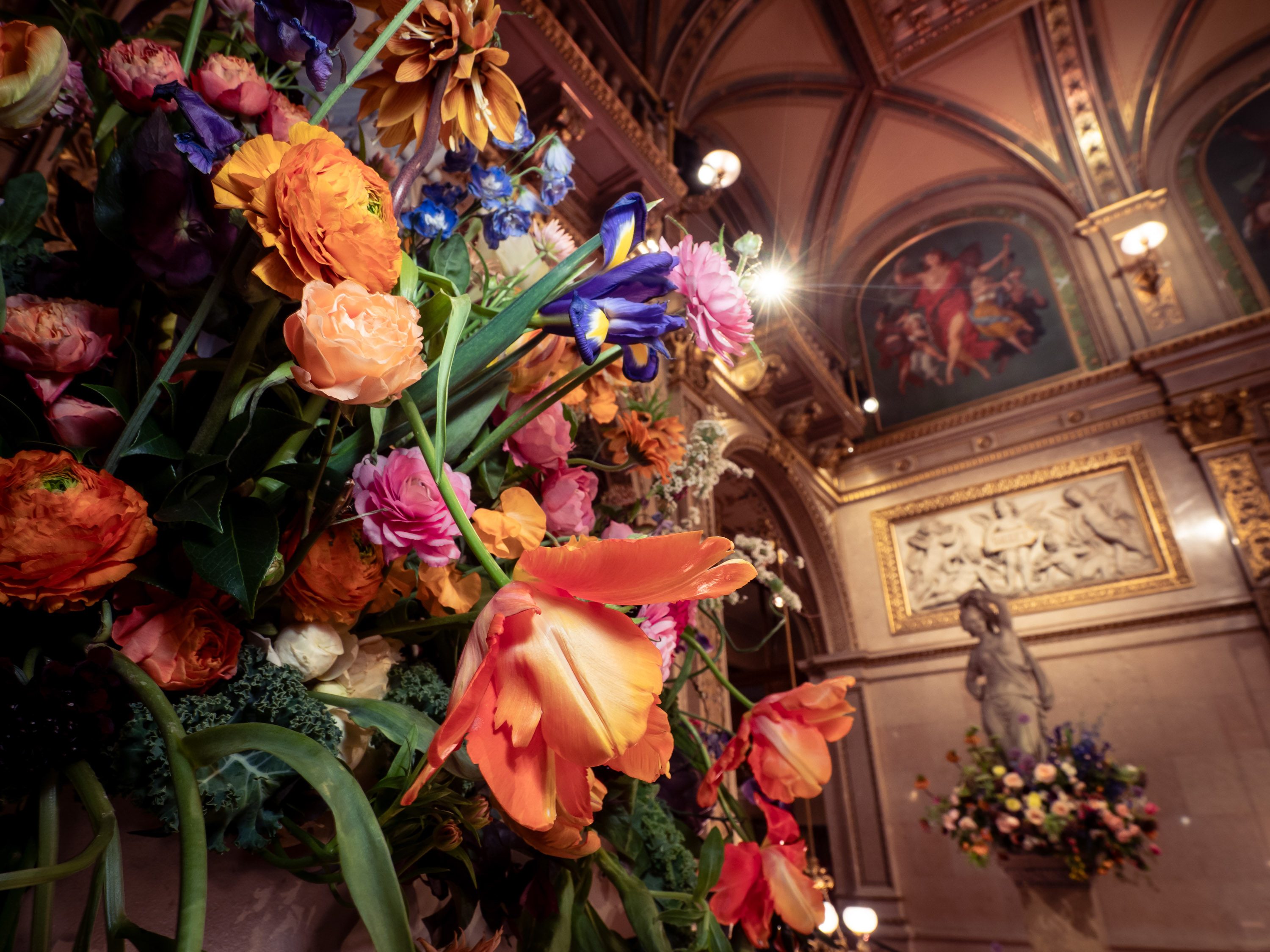 Ein farbenfrohes Blumenarrangement mit orangefarbenen Tulpen, rosa und lila Rosen sowie weiteren Blumen ist im Vordergrund zu sehen. Im Hintergrund sind kunstvoll dekorierte Wände und ein weiterer Blumenschmuck zu erkennen, der eine festliche Atmosphäre vermittelt.