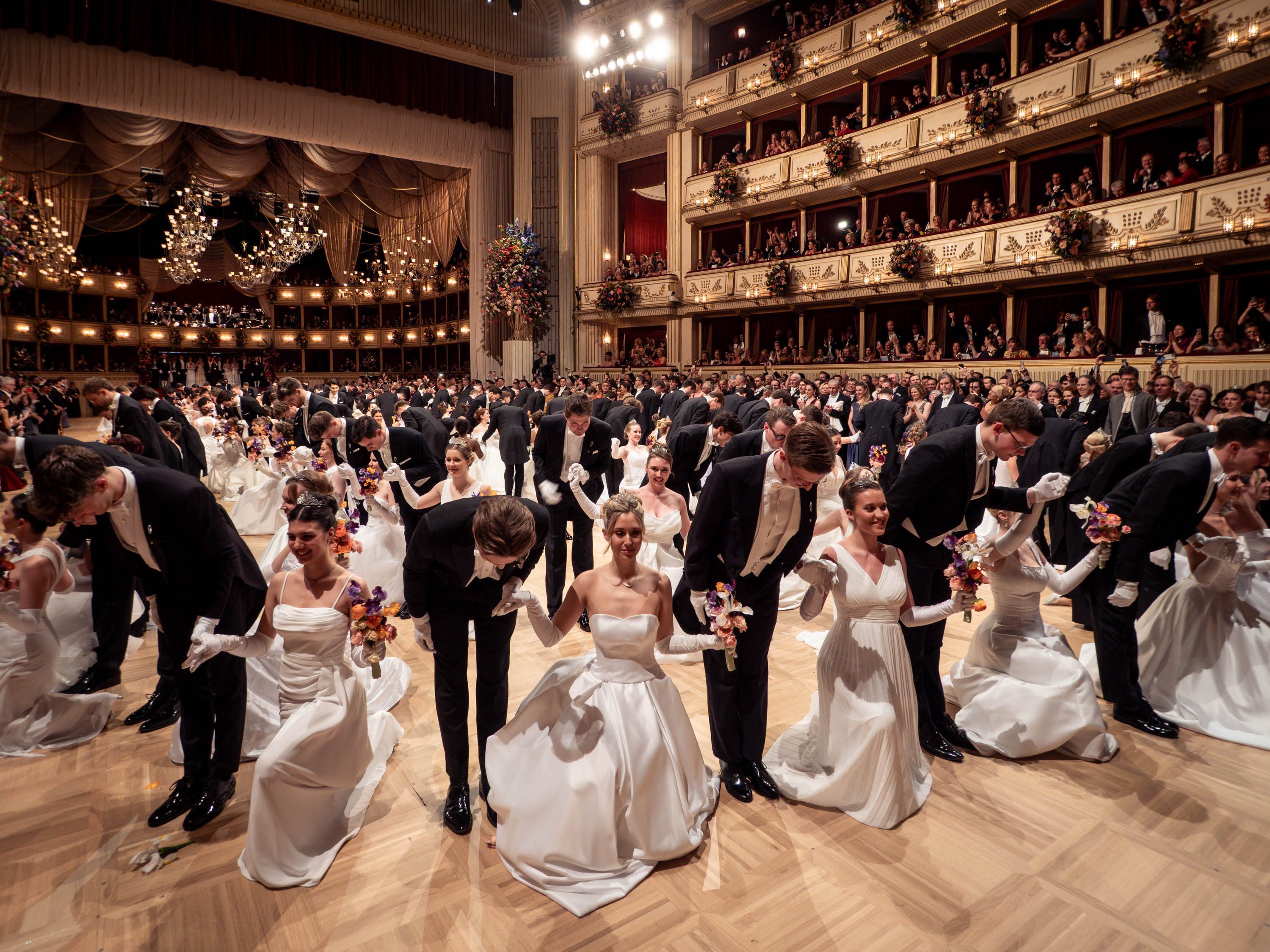 Eine große Tanzveranstaltung in einem prunkvollen Saal. Junge Paare in eleganten weißen und schwarzen Outfits knien zusammen, während sie sich verbeugen. Im Hintergrund sind Zuschauer zu sehen, die applaudieren und den Moment genießen. Der Raum ist festlich dekoriert mit Lichtern und Blumen.