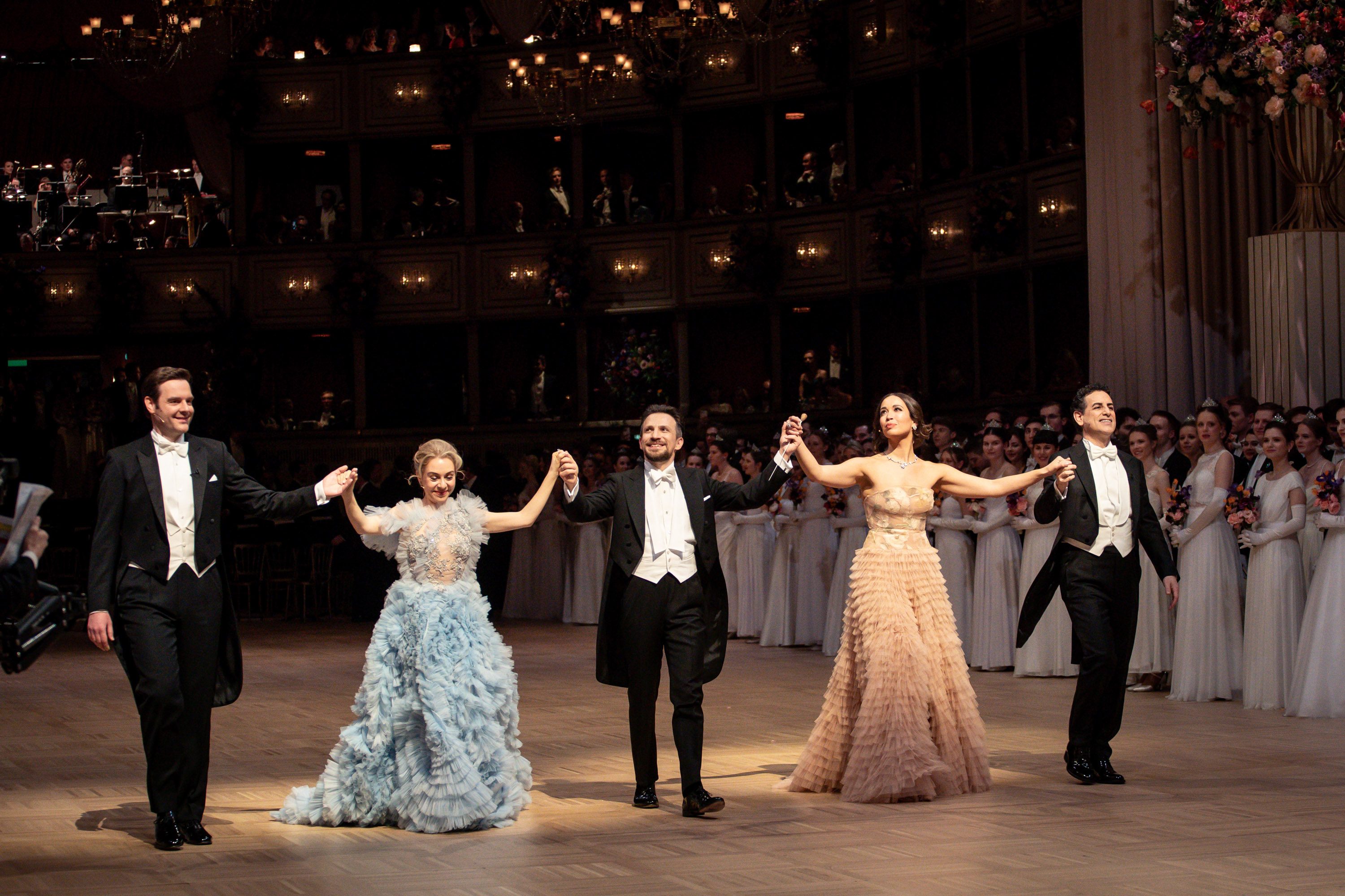 Vier elegant gekleidete Personen stehen auf einer Bühne und halten sich an den Händen. Sie lächeln in einem festlichen Ambiente, umgeben von Gästen in Abendgarderobe. Im Hintergrund sind Musiker und Zuschauer zu sehen. Es handelt sich um eine schöne Tanzaufführung oder Feier.