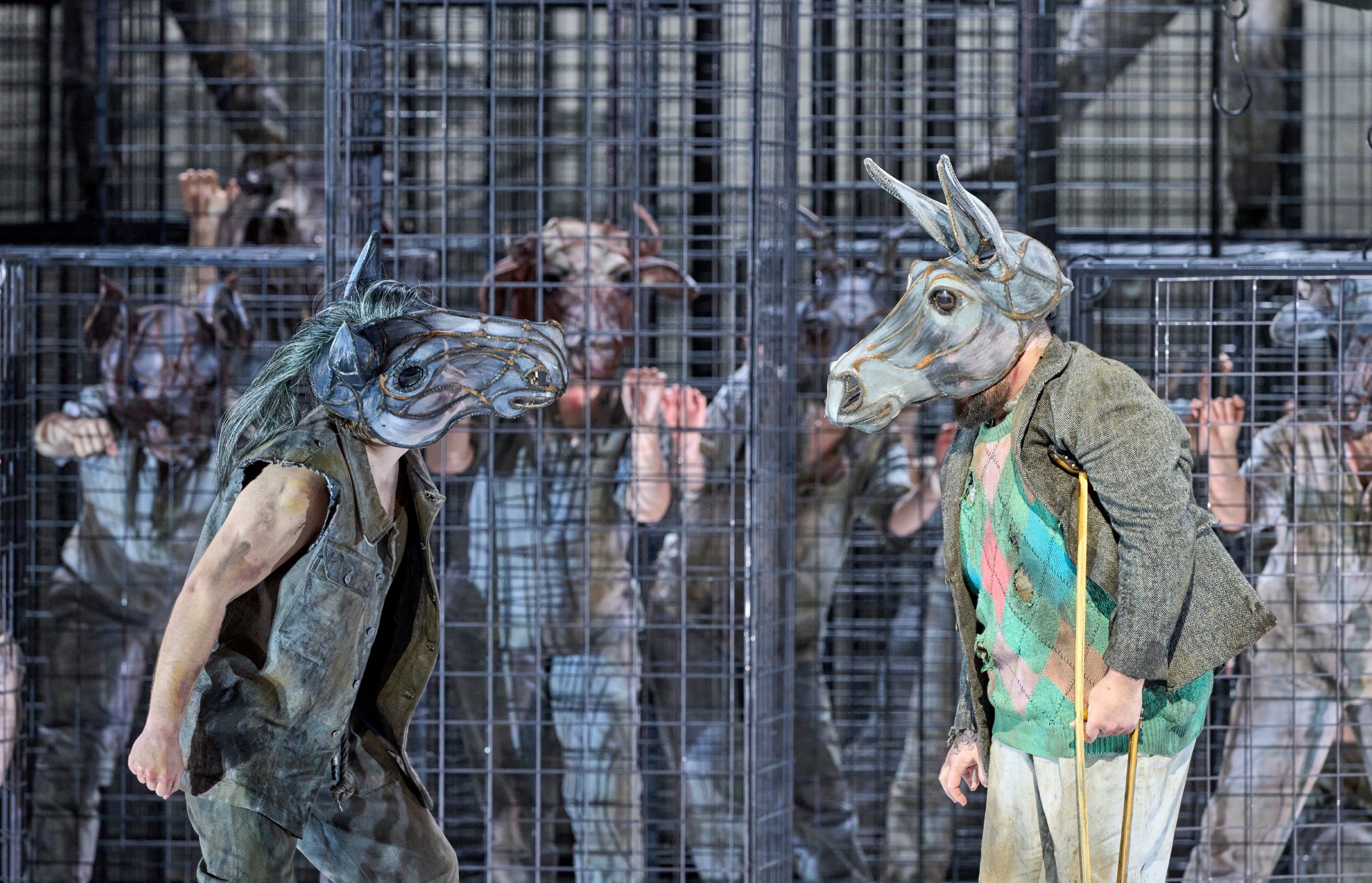 Eine Person mit einer Pferdemaske bewegt sich dynamisch vor einem Käfig, in dem mehrere Figuren mit Tiermasken gefangen sind.