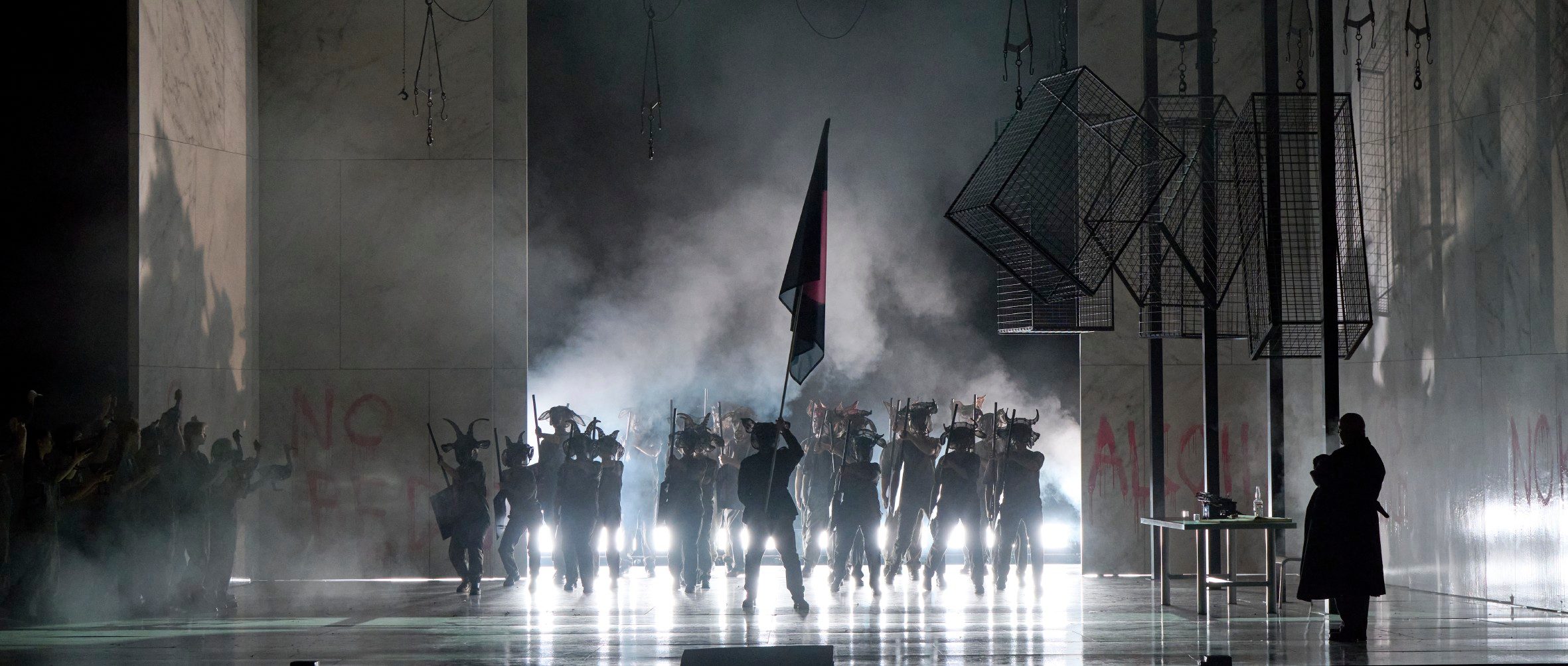 Eine Gruppe maskierter Figuren marschiert durch dichten Nebel auf einer dramatisch beleuchteten Bühne, ein Anführer trägt eine große Fahne.
