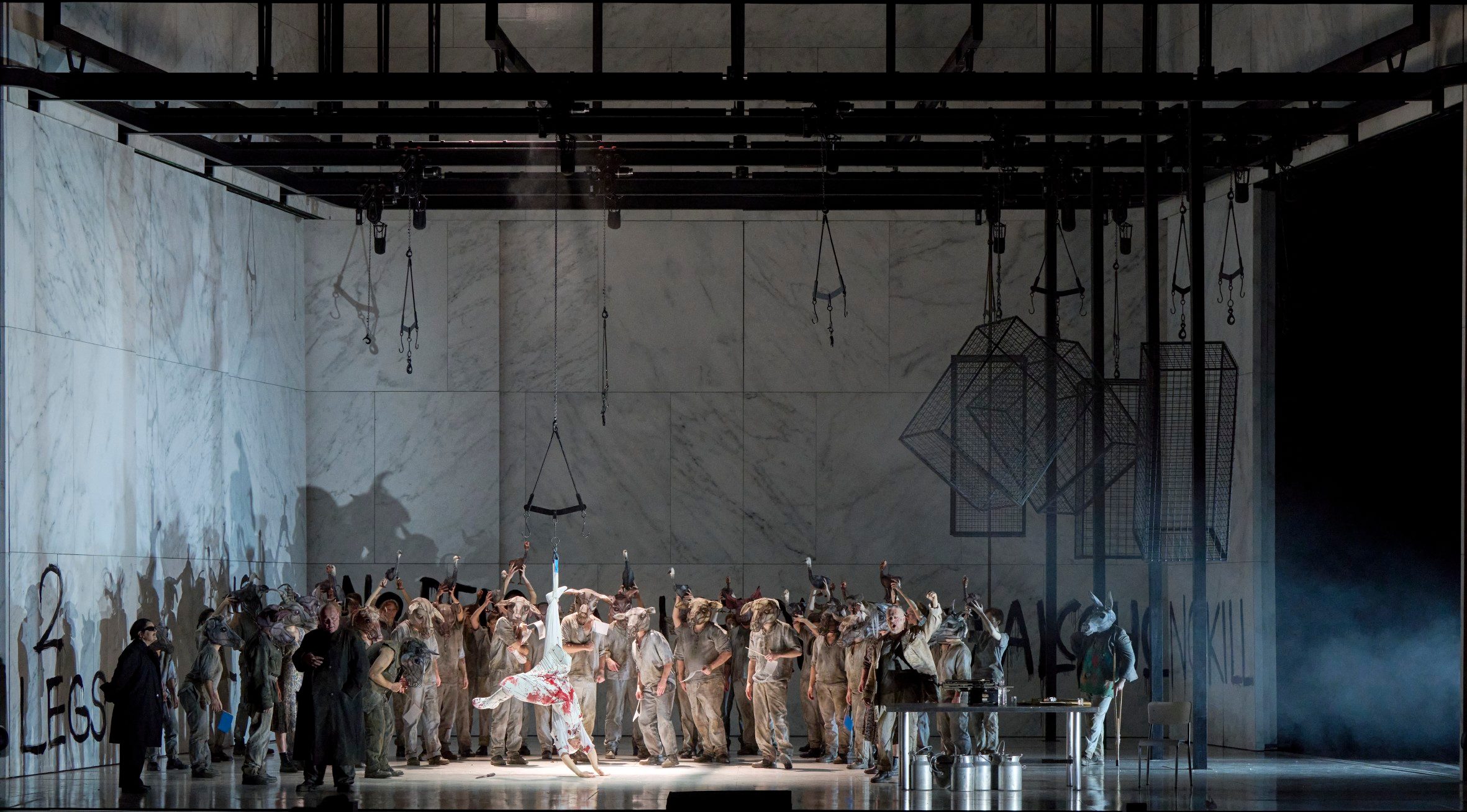 Eine große Gruppe von Menschen in grauen Kostümen umringt eine weiß gekleidete Figur mit rotem Muster, die dramatisch im Zentrum der Bühne posiert.