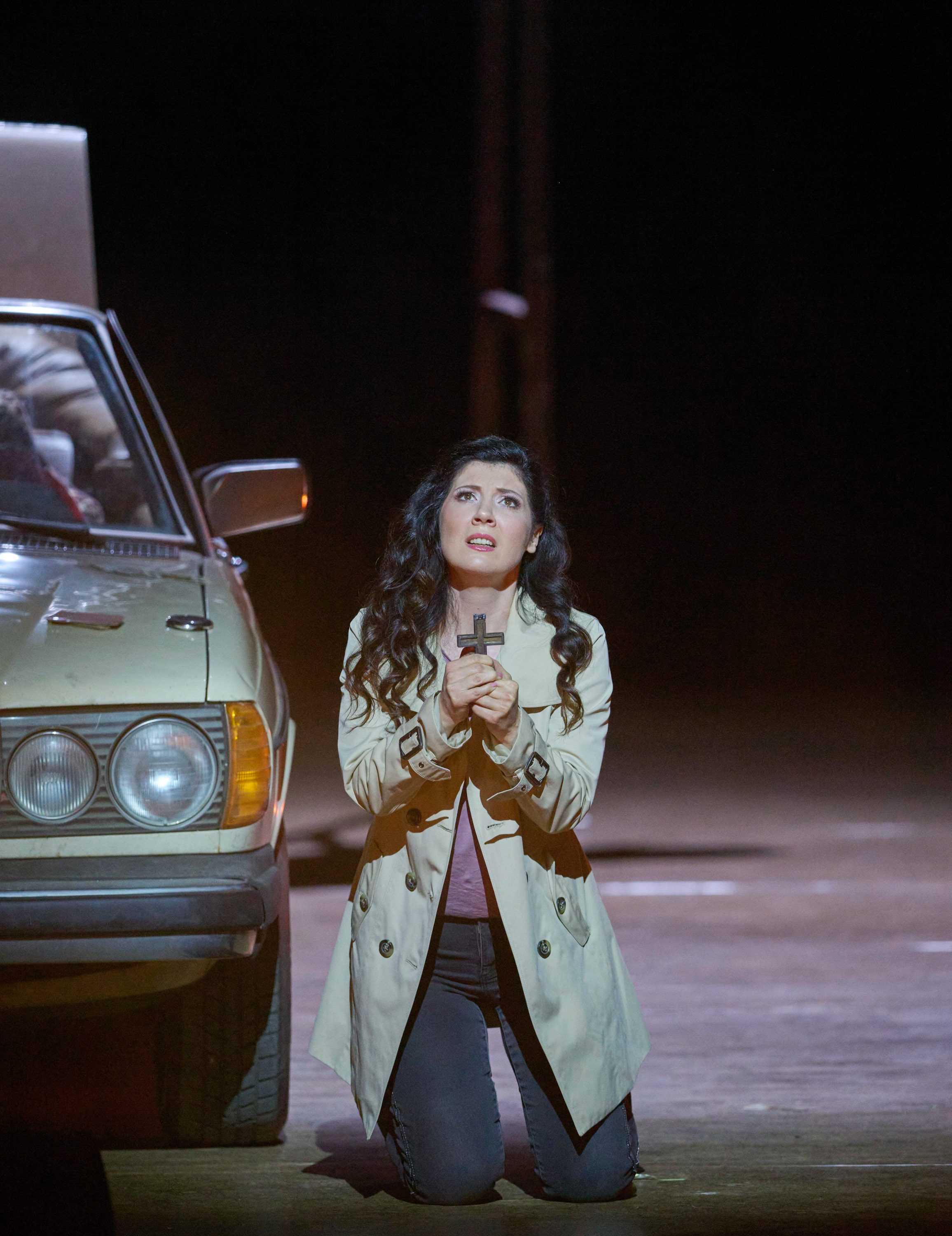 Eine Frau kniet neben einem Auto und blickt nach vorne; in den Händen hält sie ein hölzernes Kreuz