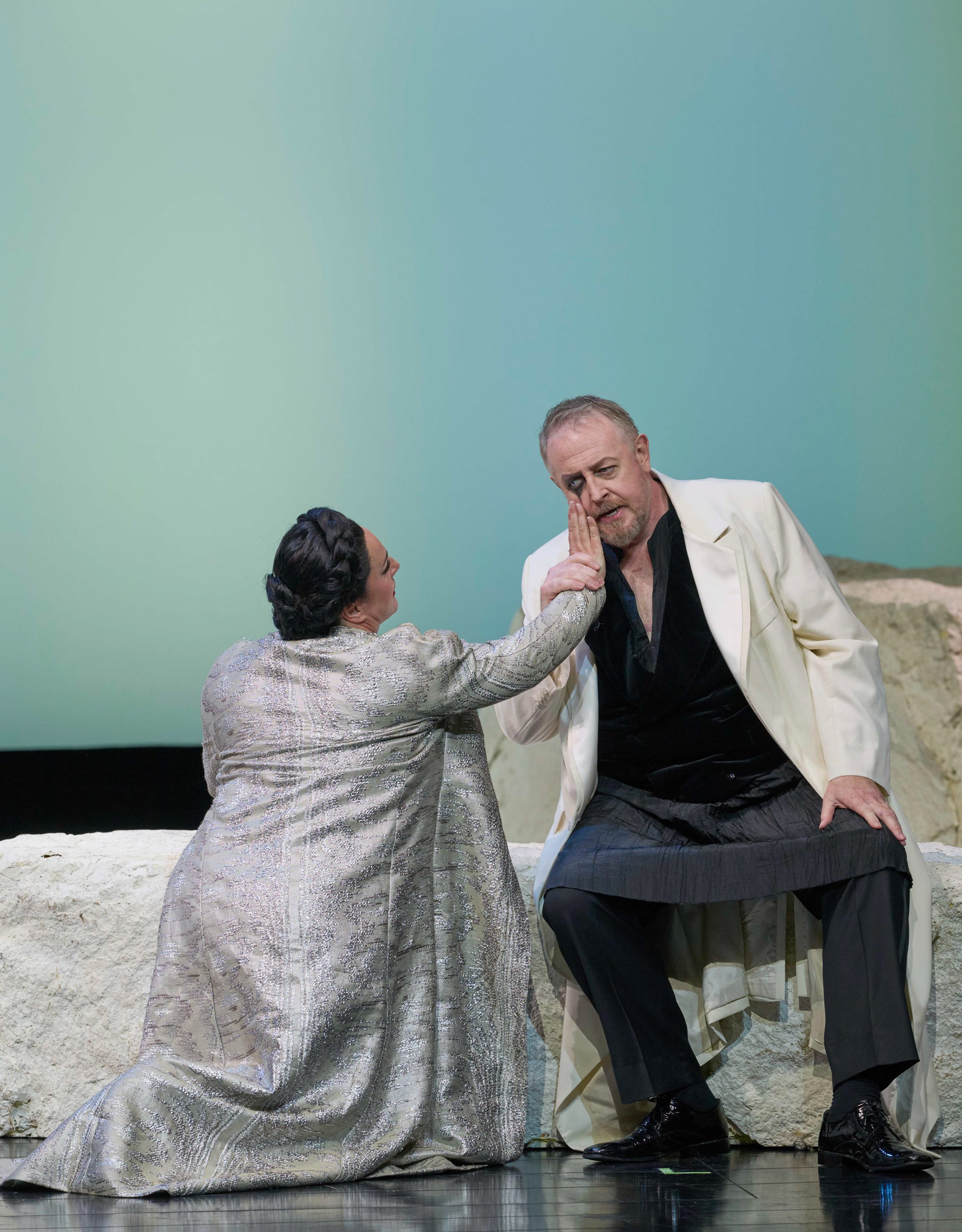 Eine Frau in einem silbernen Mantel berührt zärtlich das Gesicht eines Mannes, der auf einem Felsen sitzt.