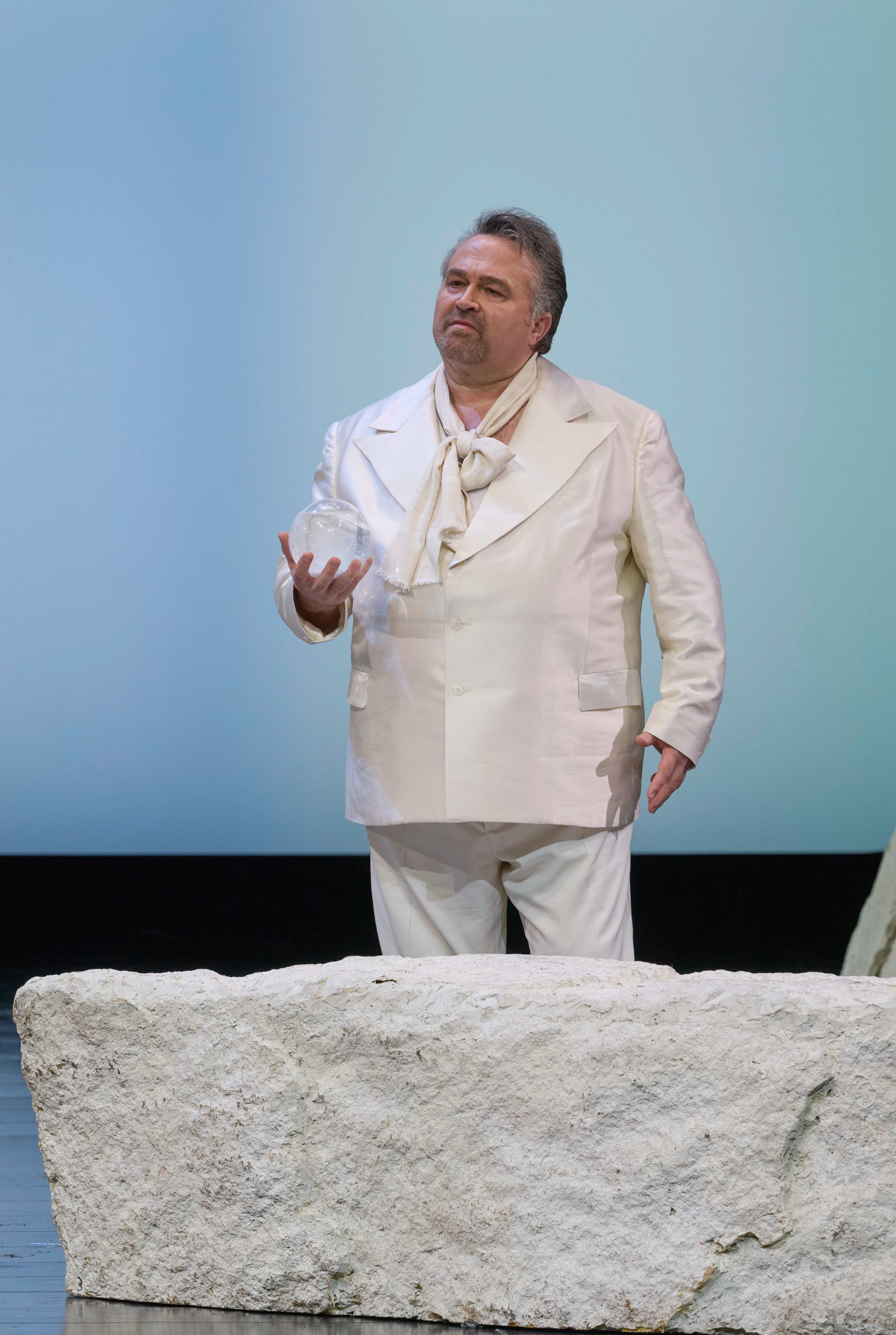 Ein Mann in einem weißen Anzug steht vor einem hellblauen Hintergrund und hält einen Glasgegenstand in der Hand.