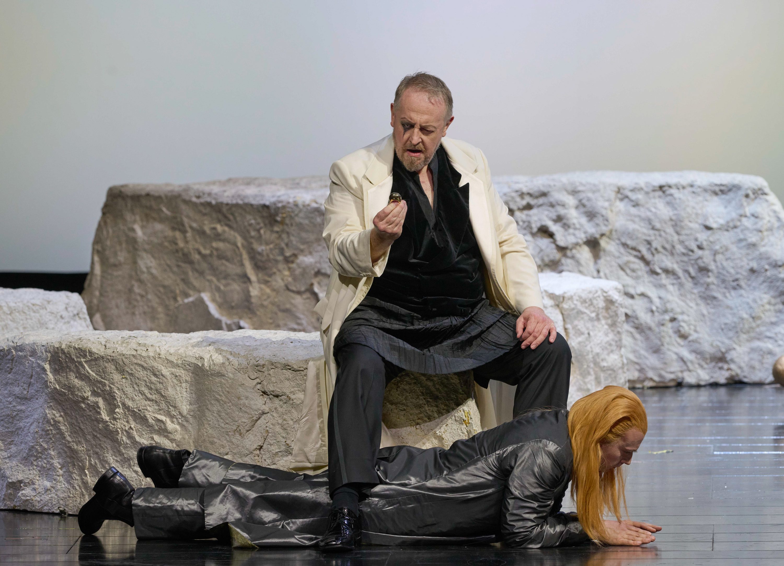 Ein Mann sitzt auf einem Felsen und beobachtet einen kleinen Gegenstand in seiner Hand; unter ihm liegt ein weiterer Mann mit langen orangenen Haaren auf dem Bauch.
