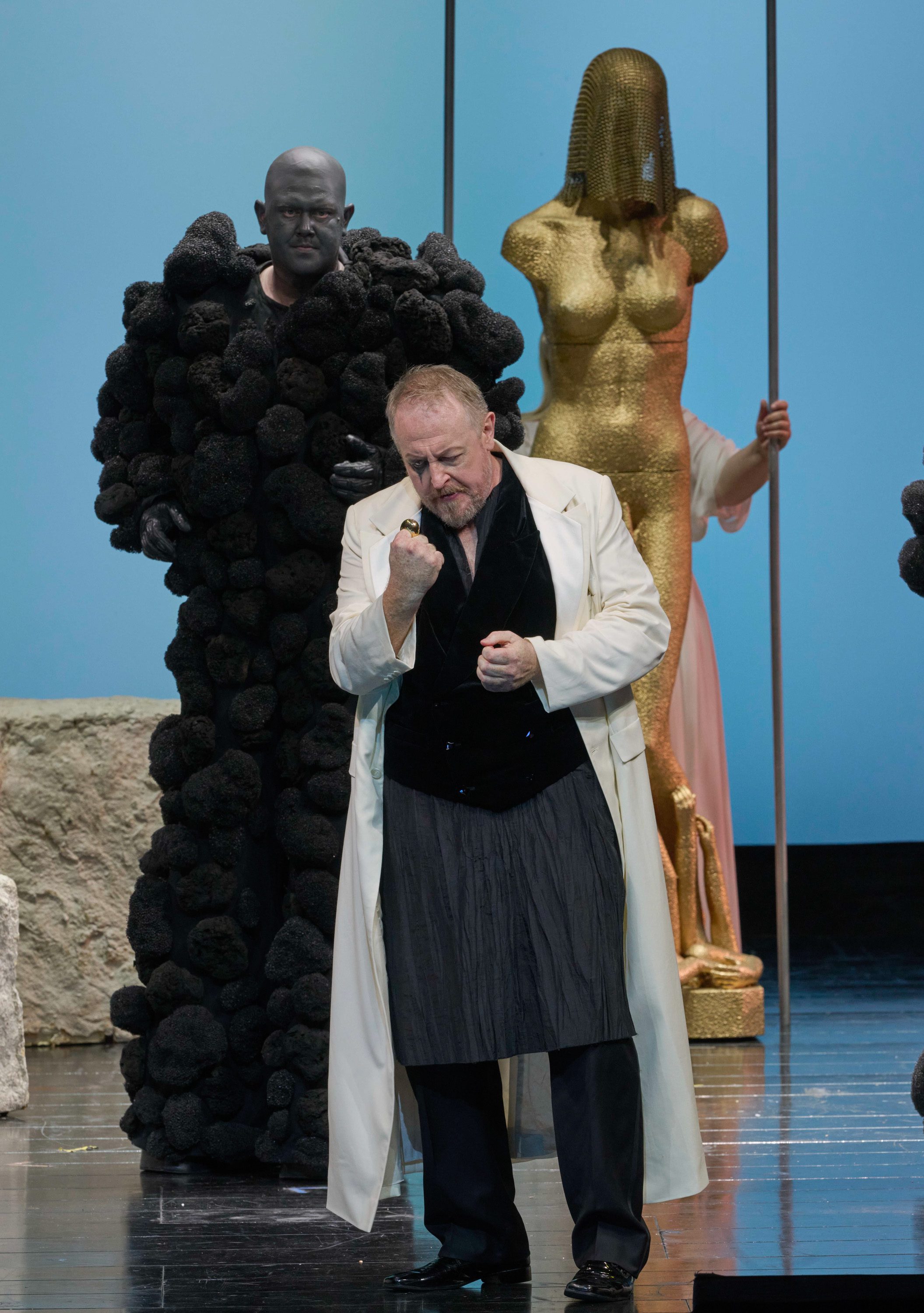 Ein Mann im weißen Mantel ballt die Faust und blickt nach unten; hinter ihm ein felsenartig dunkel kostümierter Mann.