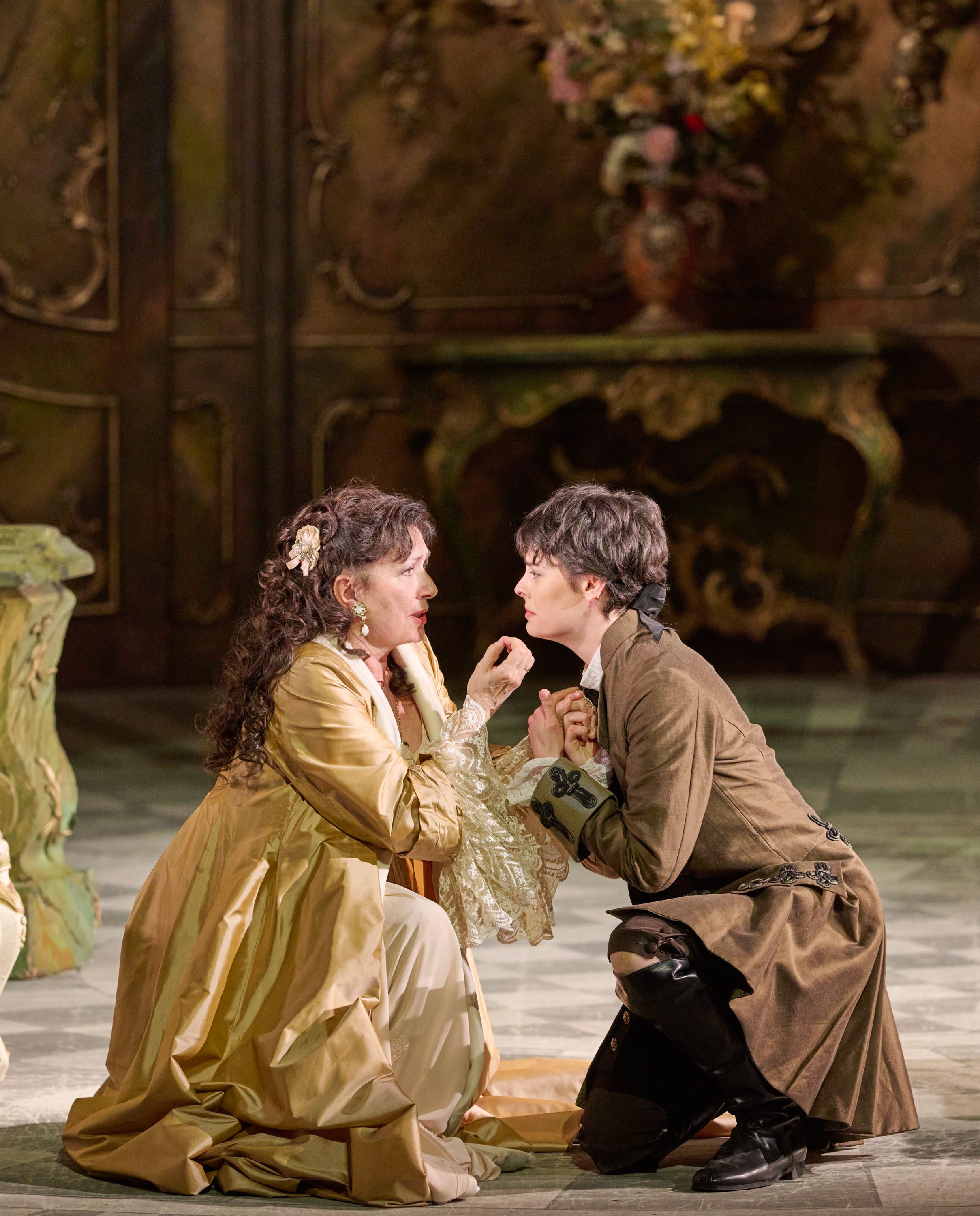 Zwei Schauspielerinnen in historischer Kostümierung interagieren intensiv auf einer Bühne. Eine Person im goldenen Kleid schaut die andere, die in brauner Kleidung sitzt, eindringlich an, während sie ihre Hände hält. Der Hintergrund zeigt eine dekorative Kulisse mit Pflanzen.