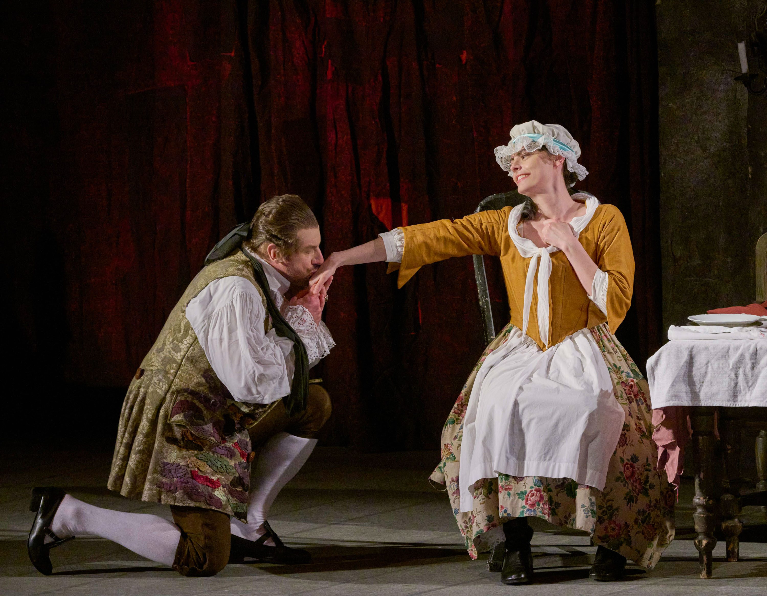 Ein Mann kniet vor einer Frau und küsst sanft ihre Hand. Die Frau, in einem historischen Kleid, lächelt. Im Hintergrund sind schwach beleuchtete Wände und ein Tisch mit Geschirr sichtbar. Die Szene vermittelt einen Moment romantischer Zuneigung.