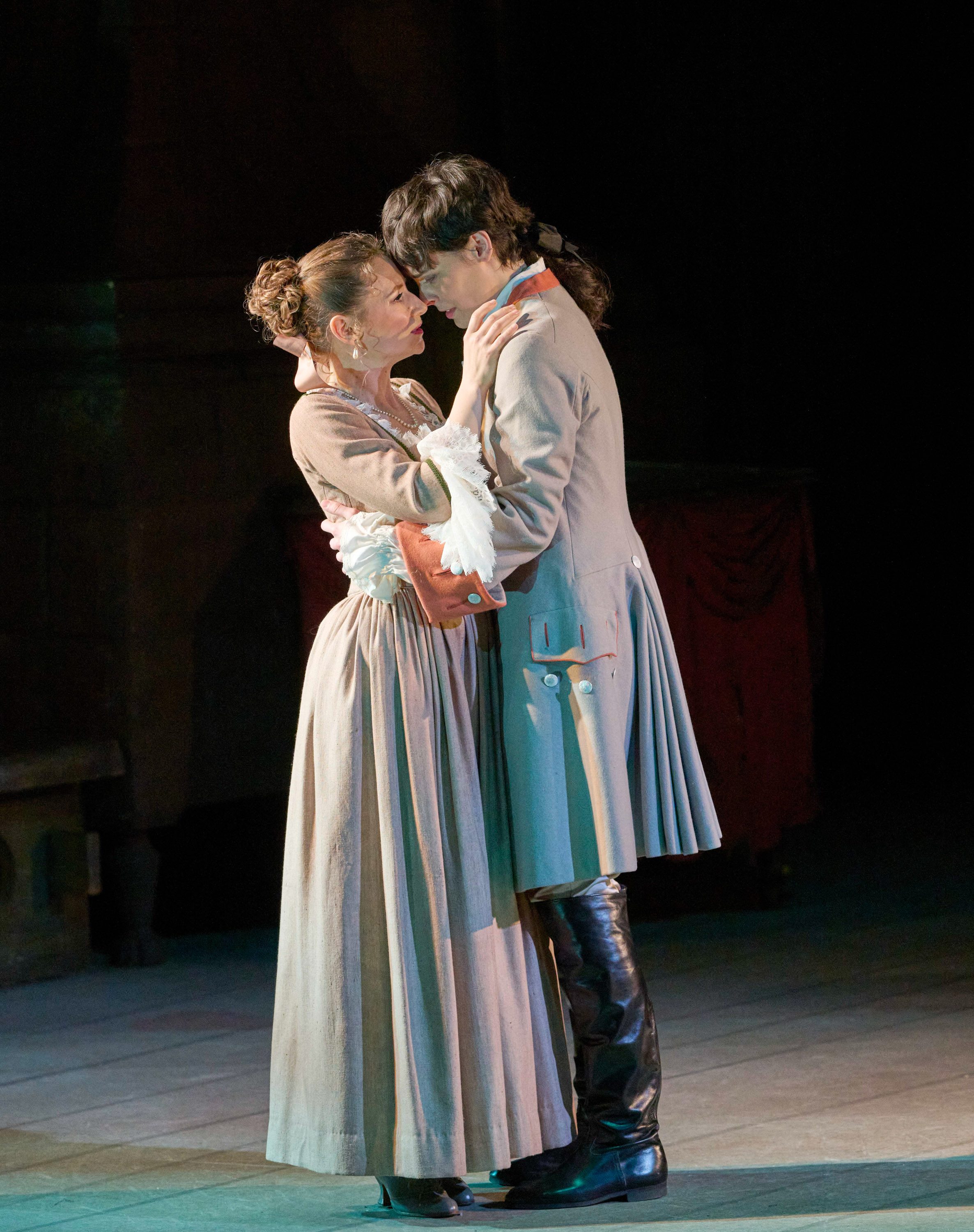 Zwei Figuren stehen sich gegenüber und umarmen sich zärtlich. Sie sind historisch gekleidet, mit einem langen Kleid und einem Anzug. Ihre Gesichter sind nahe beieinander, was eine intime Verbindung zwischen ihnen zeigt. Der Hintergrund ist dunkel, wodurch die beiden zentralen Figuren hervorgehoben werden.