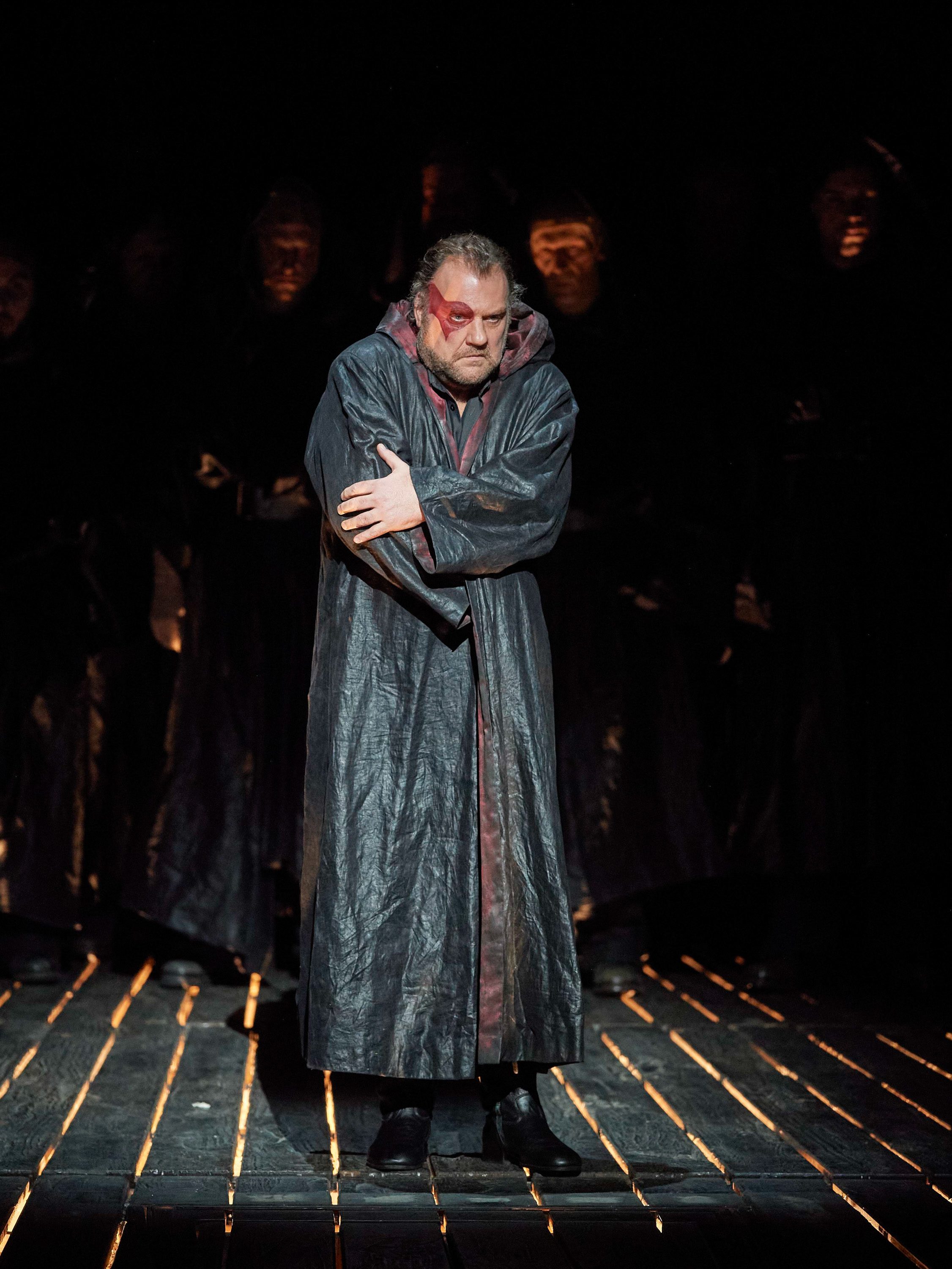 Ein verletzter Mann in dunklem Gewand steht im Scheinwerferlicht vor einer Gruppe schwarz gekleideter, maskierter Gestalten.