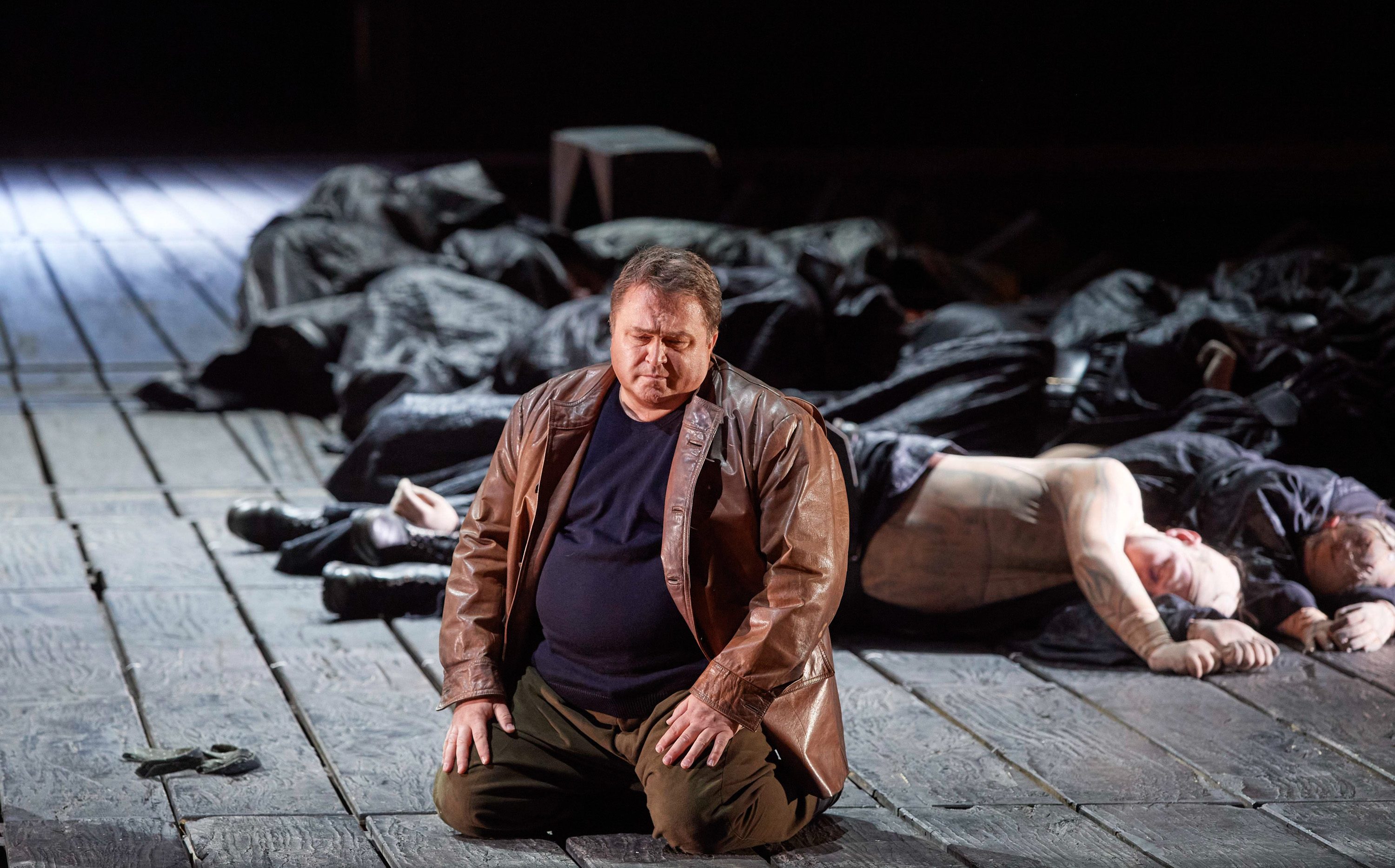 Ein Mann in Lederjacke kniet auf einem Holzboden und blickt verzweifelt nach vorne..