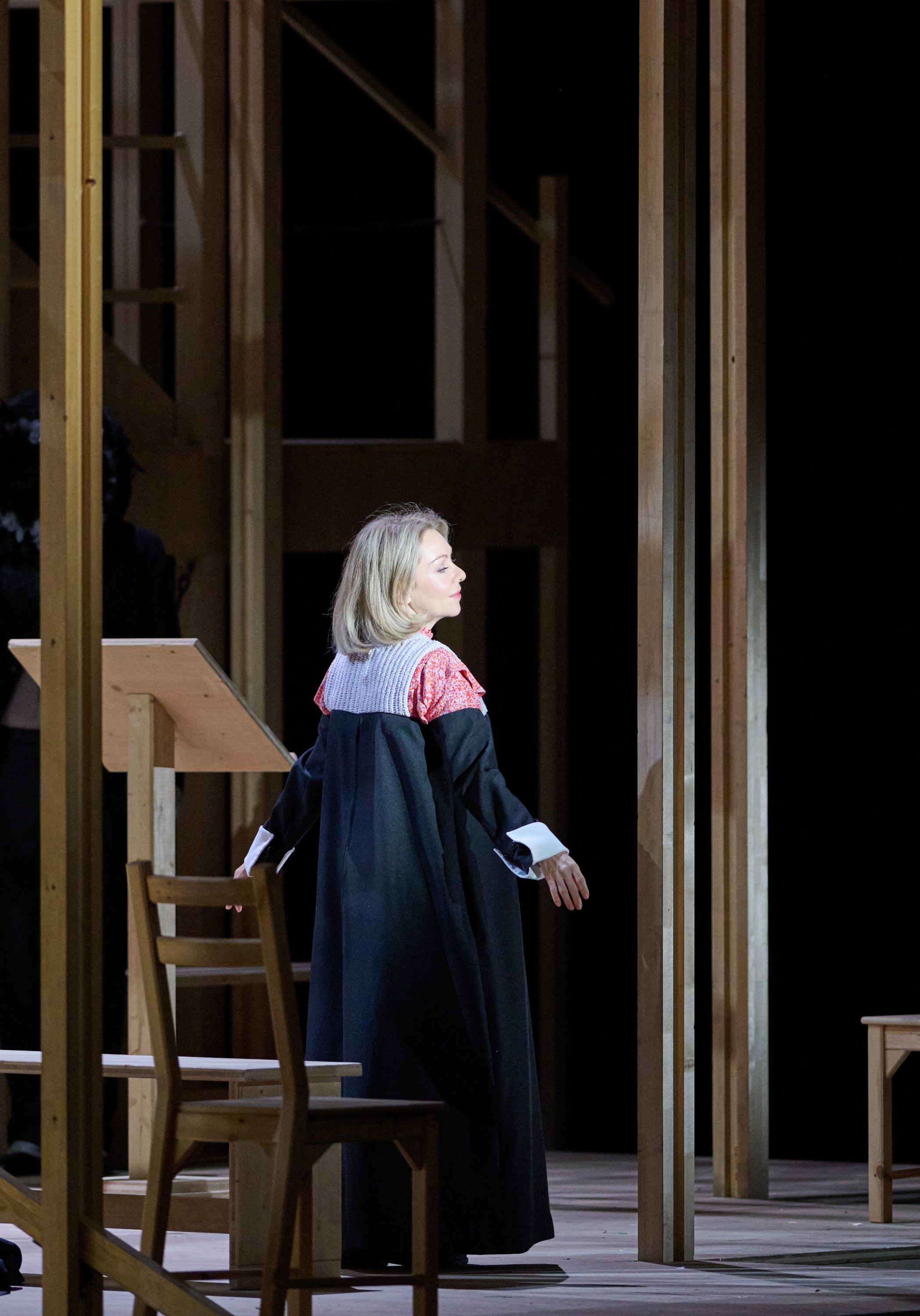 A woman in a black robe with a red and gray collar is standing on stage, facing away and looking to her side. In the background, wooden structures and a wooden lectern are visible, suggesting a theatrical setting.