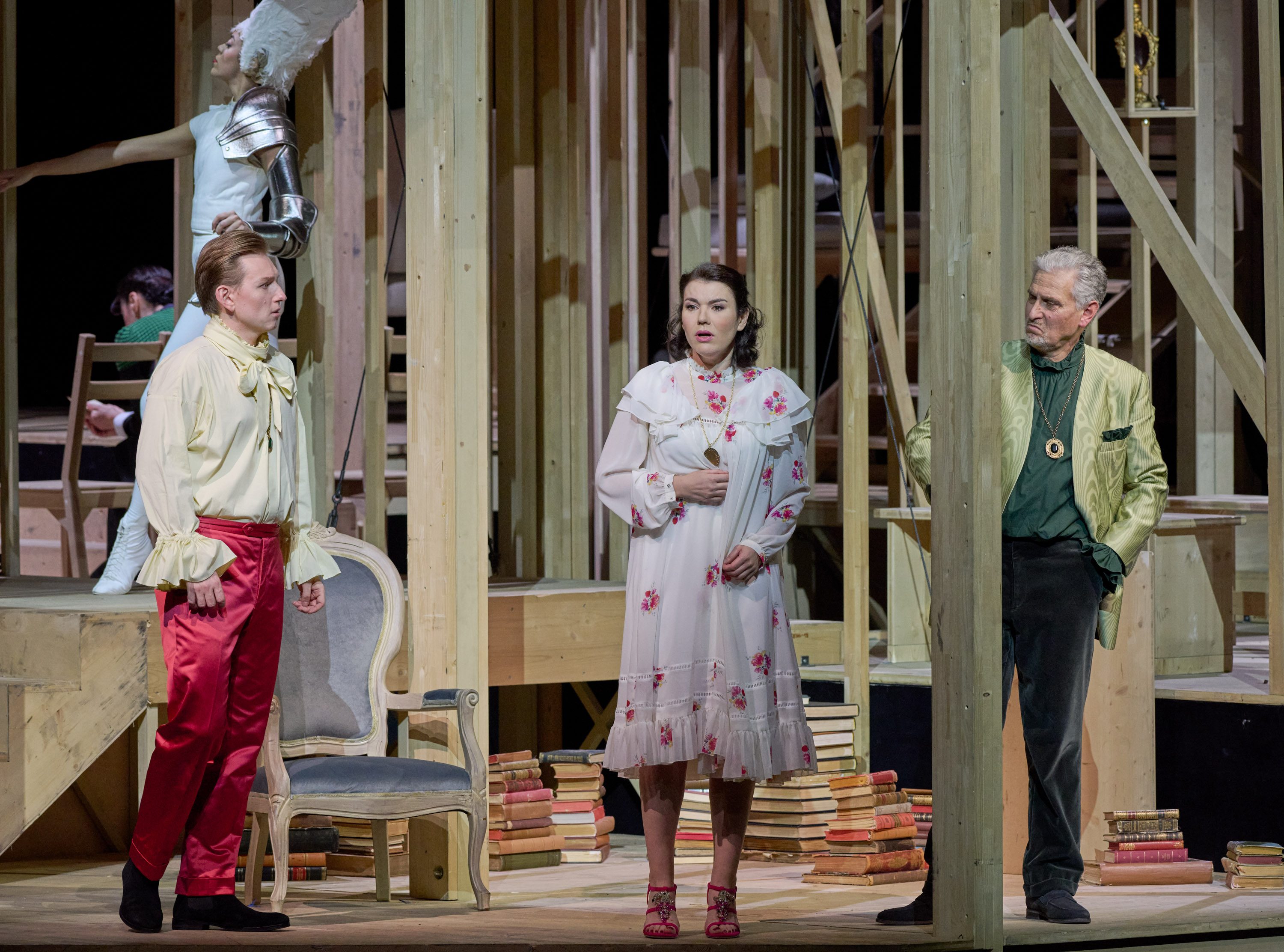 A theatrical scene features three characters: a man in a yellow shirt and red pants, a woman in a white dress with floral patterns, and an older man in a green shirt and gold jacket. They stand on a wooden stage with stacks of books nearby, suggesting a dramatic moment.