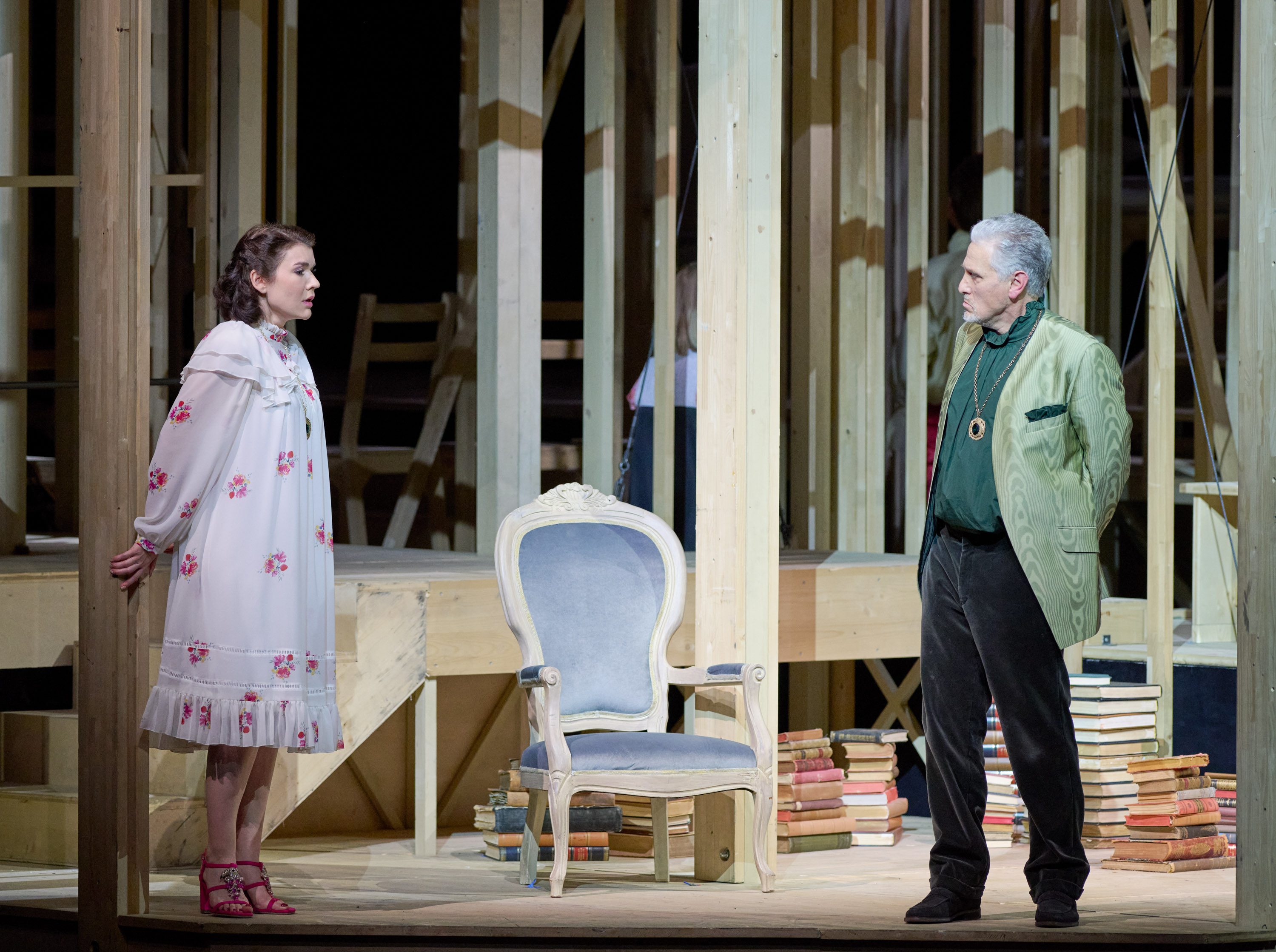 A scene from a theatrical performance features two actors engaged in conversation. One is a woman in a floral white dress, standing near a wooden pillar, while the other is a man in a green jacket, standing with his hands in his pockets. A blue chair and stacked books are visible nearby.