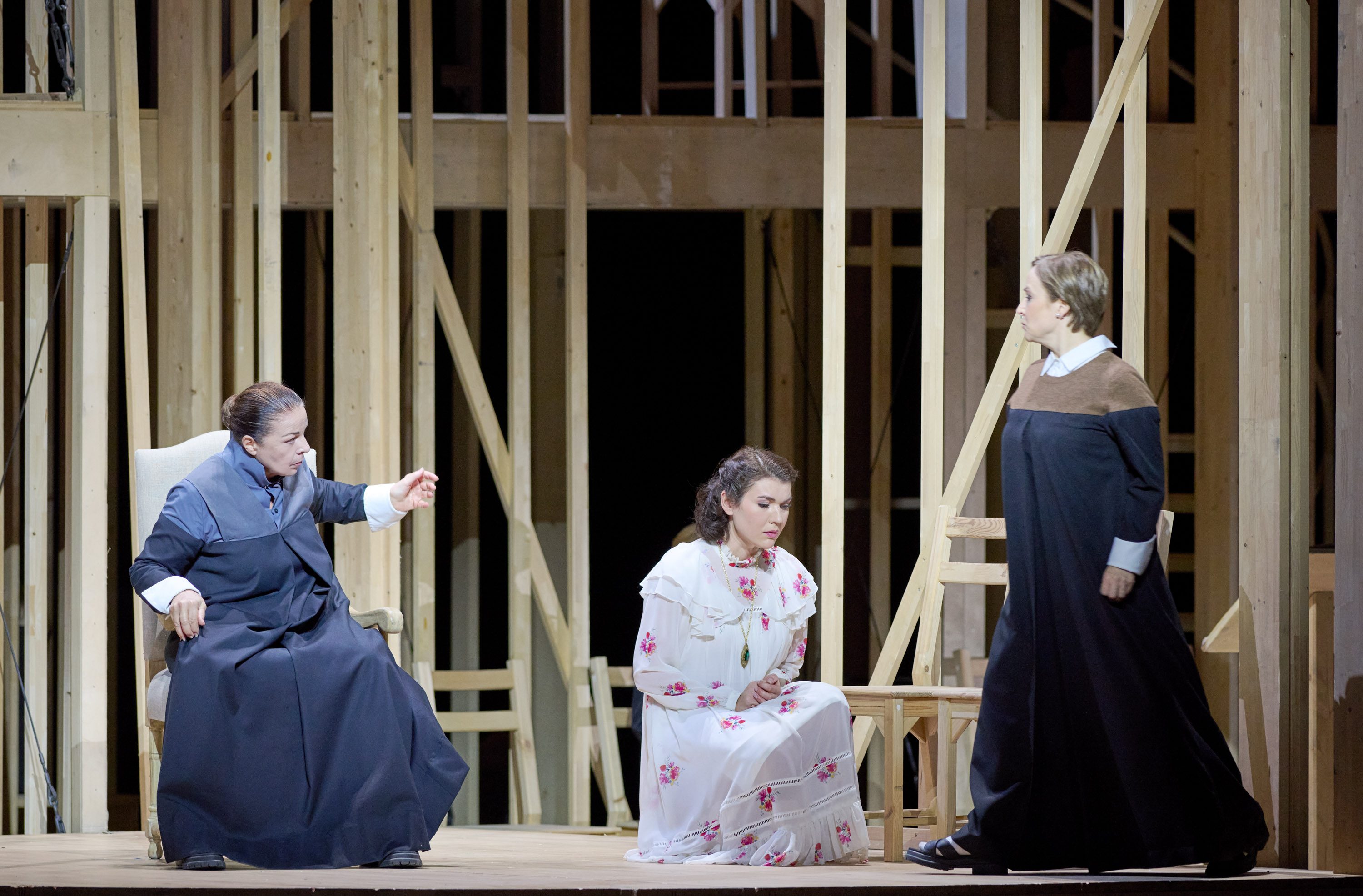 Three women are depicted in a dramatic scene. One woman sits on a chair, gesturing animatedly, while another kneels on the floor, looking downcast. The third woman stands nearby, observing. The background features wooden stage framing, suggesting a theatrical setting.