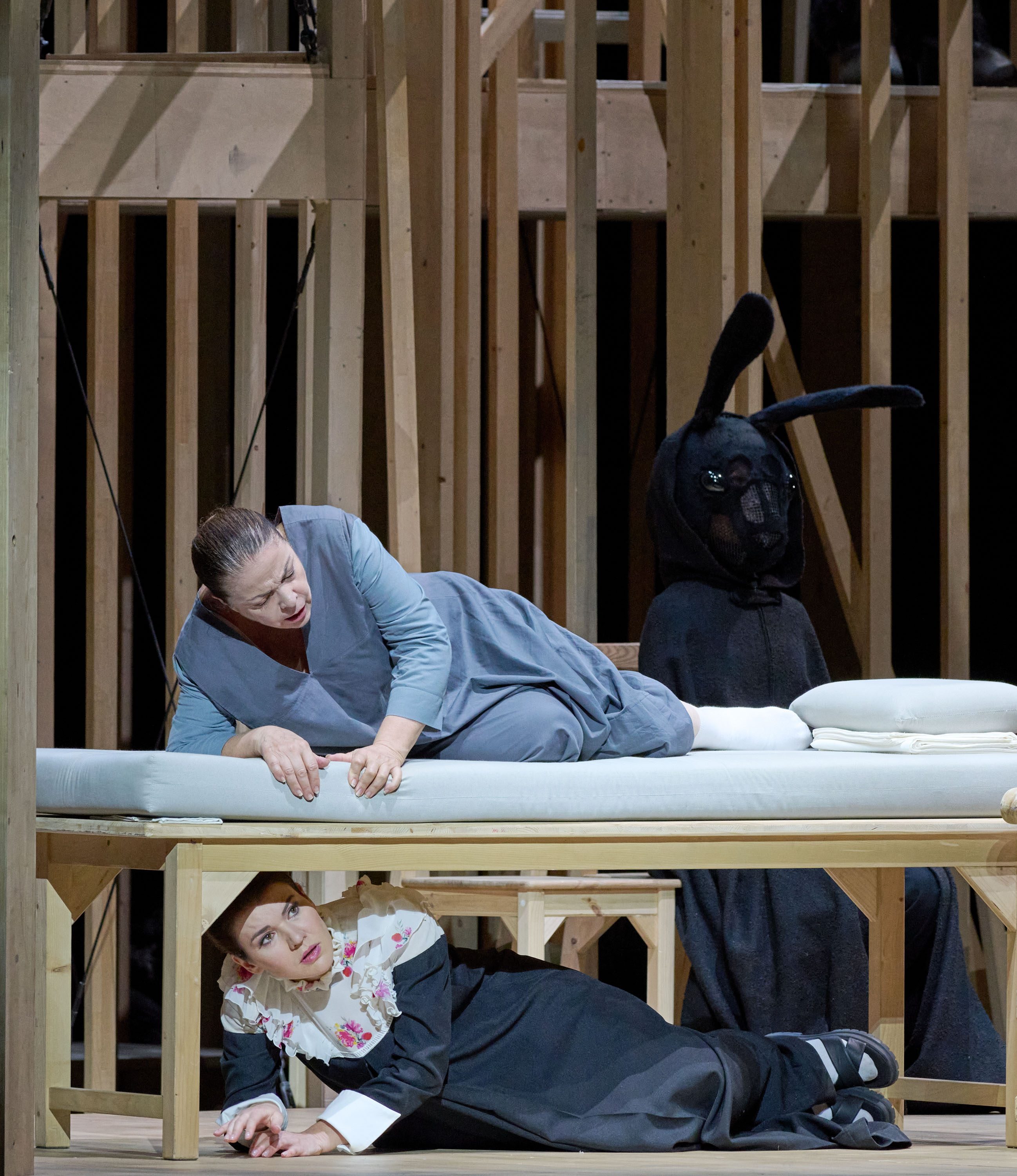 A theatrical scene shows two women in dramatic poses. One woman, in a gray dress, is leaning over a bed, while another woman, dressed in black with floral accents, is crawling underneath the bed. A figure in a dark rabbit costume observes in the background, adding an eerie element.