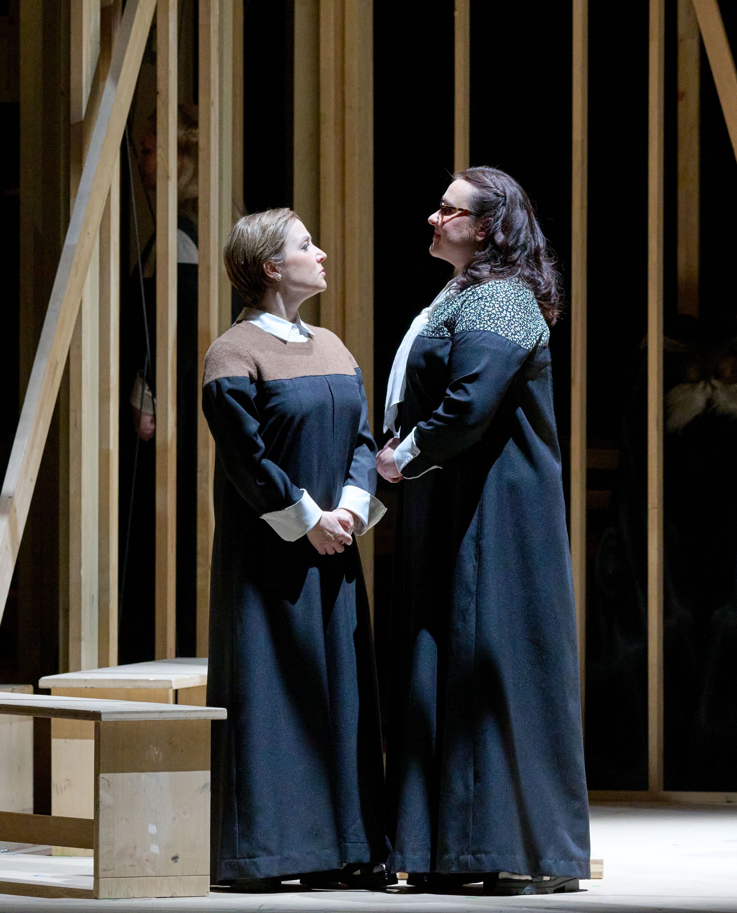 Two women in long black dresses stand in a spotlight, facing each other with expressions of intensity. One woman has short hair and a brown-sleeved garment, while the other has long hair with a patterned collar. They are surrounded by a wooden frame set, suggesting a theatrical performance.