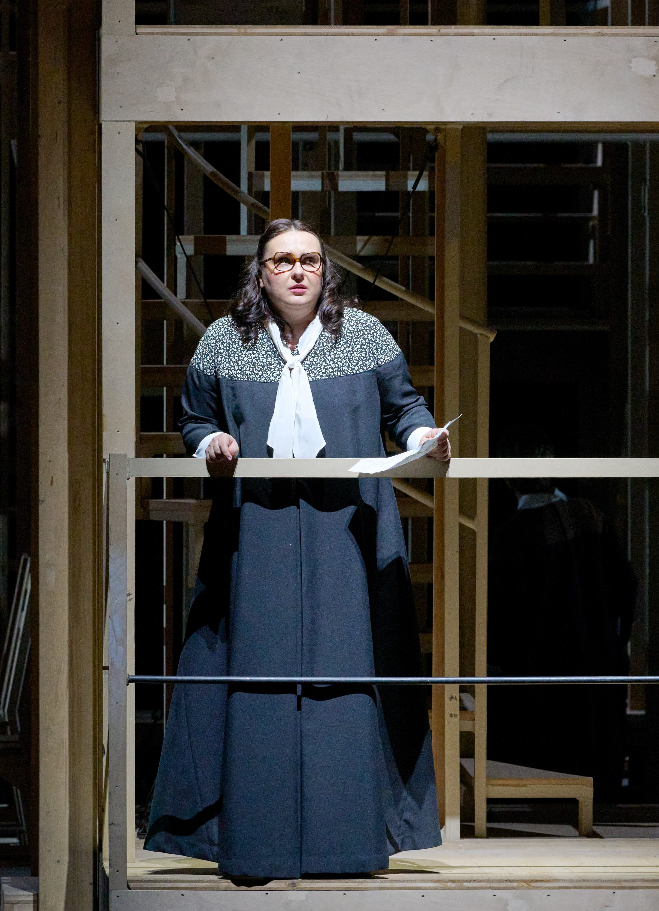 A performer stands on a wooden platform, singing passionately. She wears a long black dress with a patterned neckline and a white bow tie. The backdrop features wooden structures, suggesting a theatrical setting. Her expression is focused, conveying emotion as she holds a script in one hand.